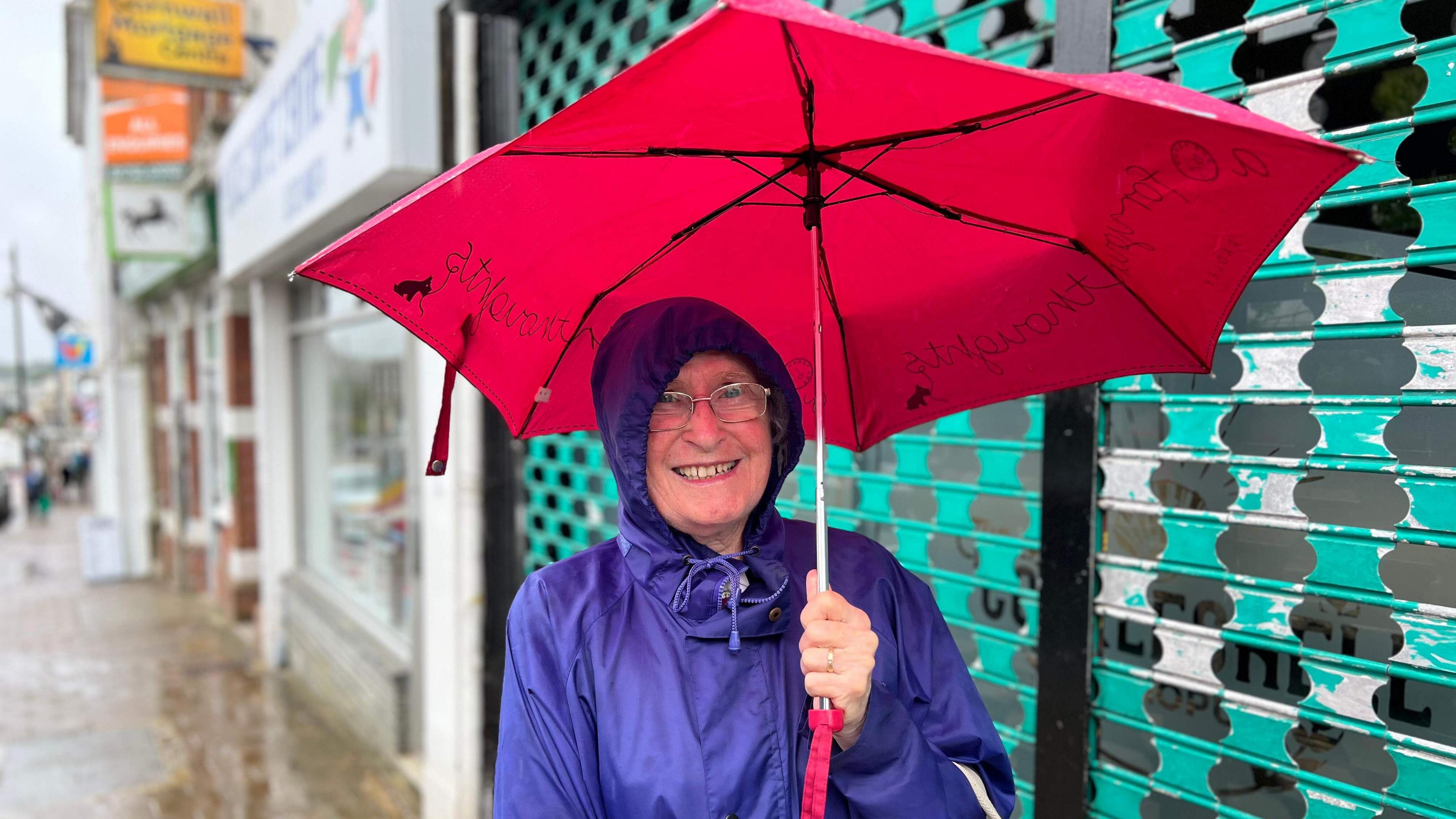 A very smiley lady in purple raincoat with hood tightened around her face holds a red umbrella over her head while smiling at the camera standing in front of shops on Saltash High Street