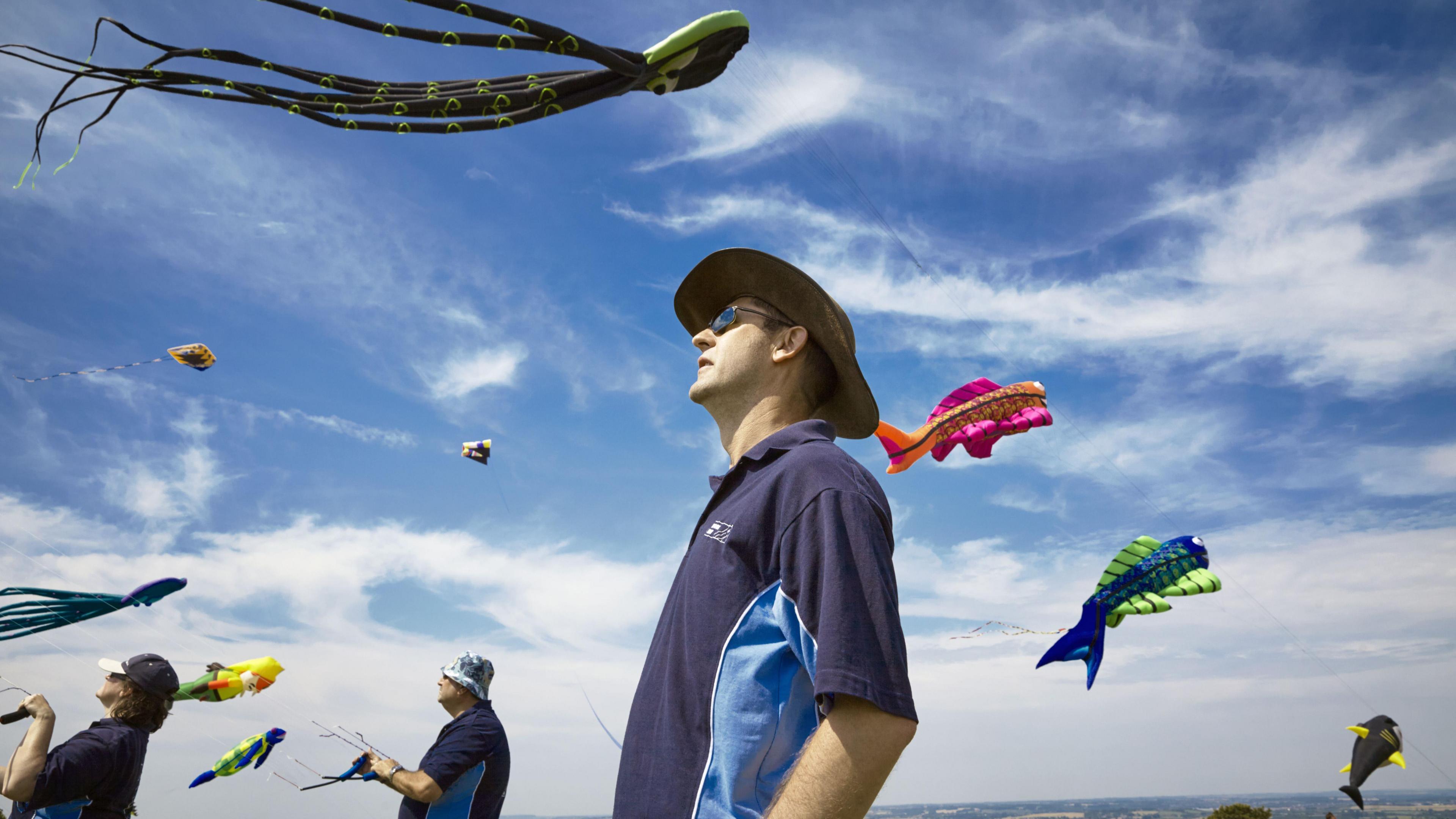 A man in a blue top, wearing sunglasses and a hat, looking at kites taking off, with two other people flying kites in the distance