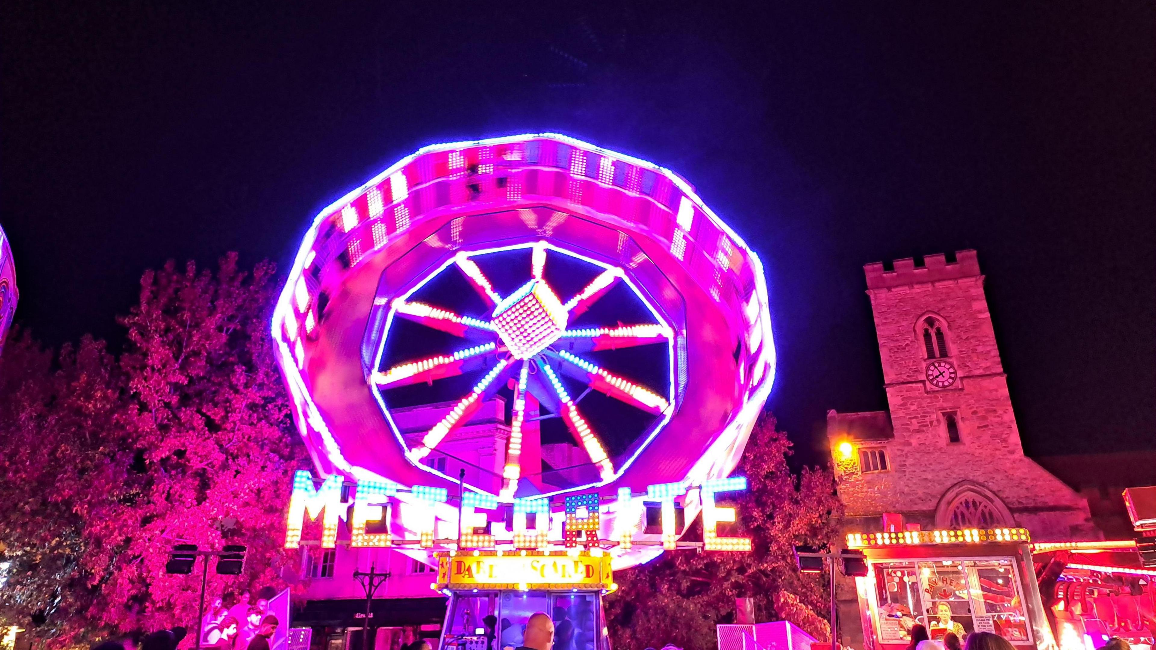 A spinning funfair ride glows bright neon pink against a night sky. It's light is illuminating a church tower and trees in background.