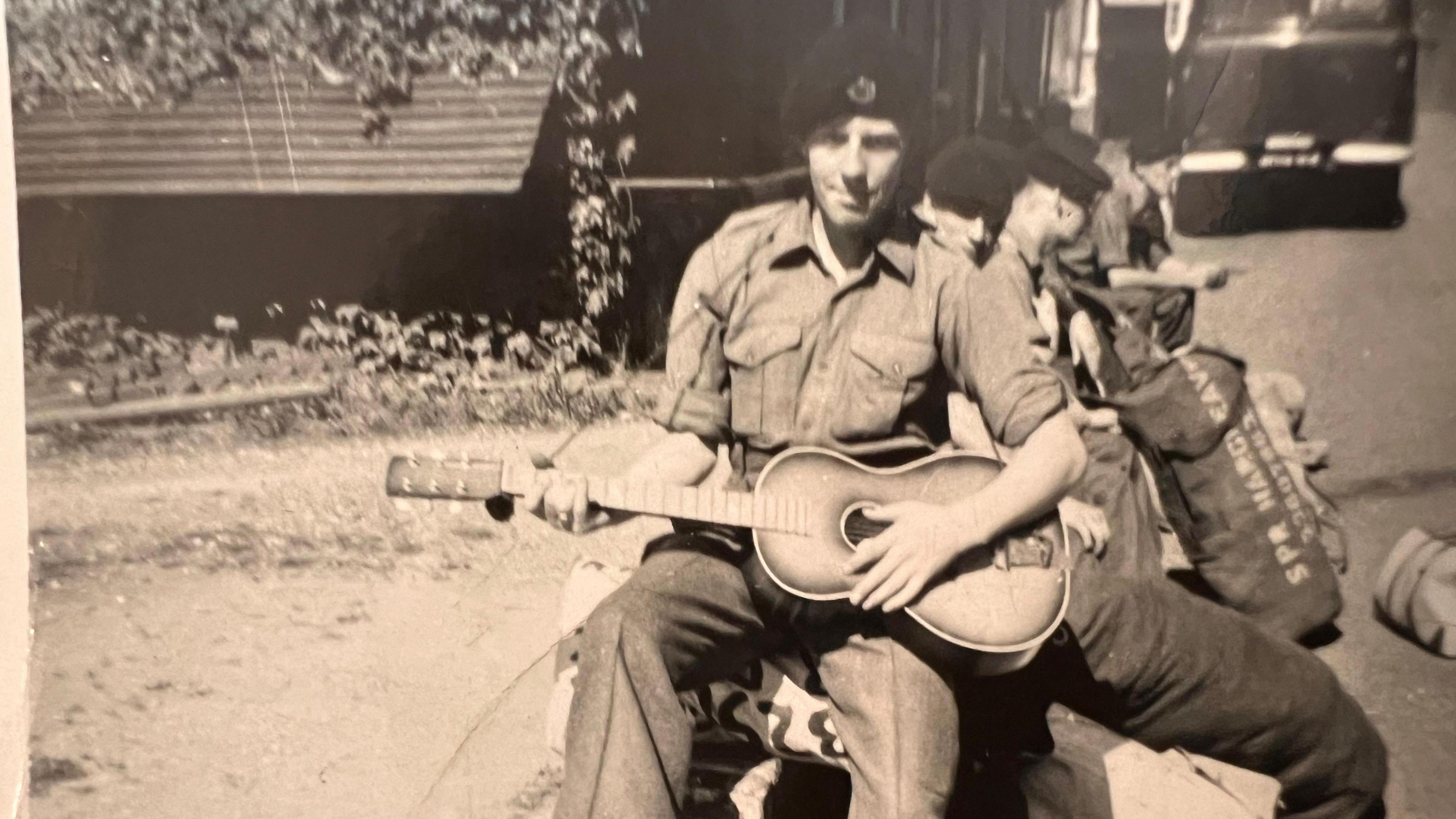 Mr Peace holds a guitar and looks at the camera. He is wearing a shirt and trousers and a black beret-style cap in the black and white photo.