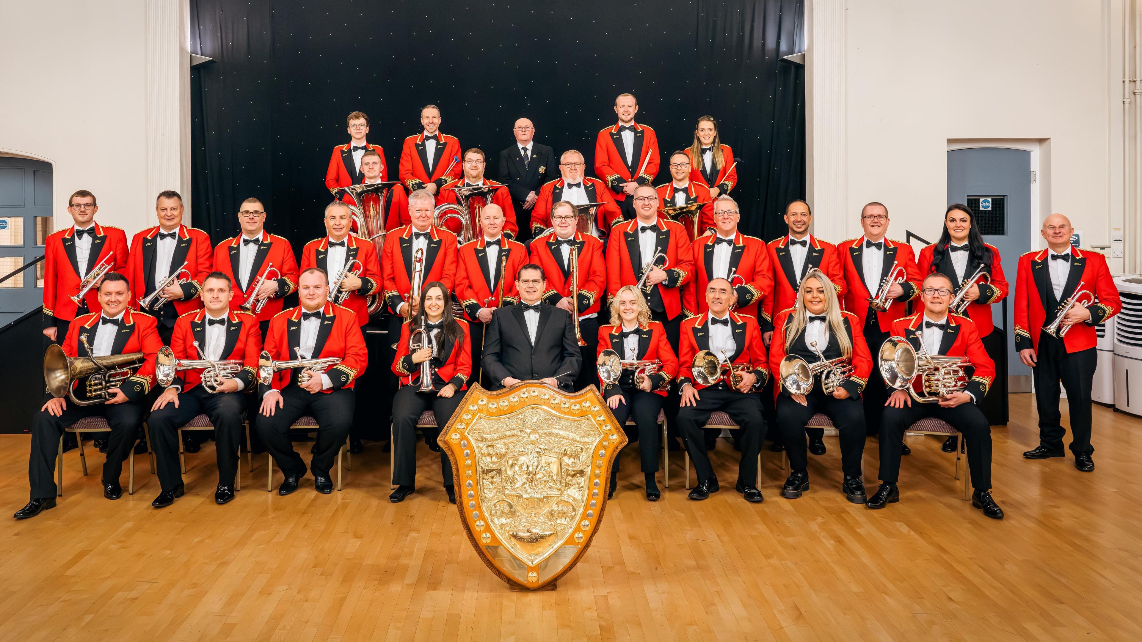 Members of a brass band wearing red jackets, bow ties and holding instruments, sit behind a big golden shield in a town hall.