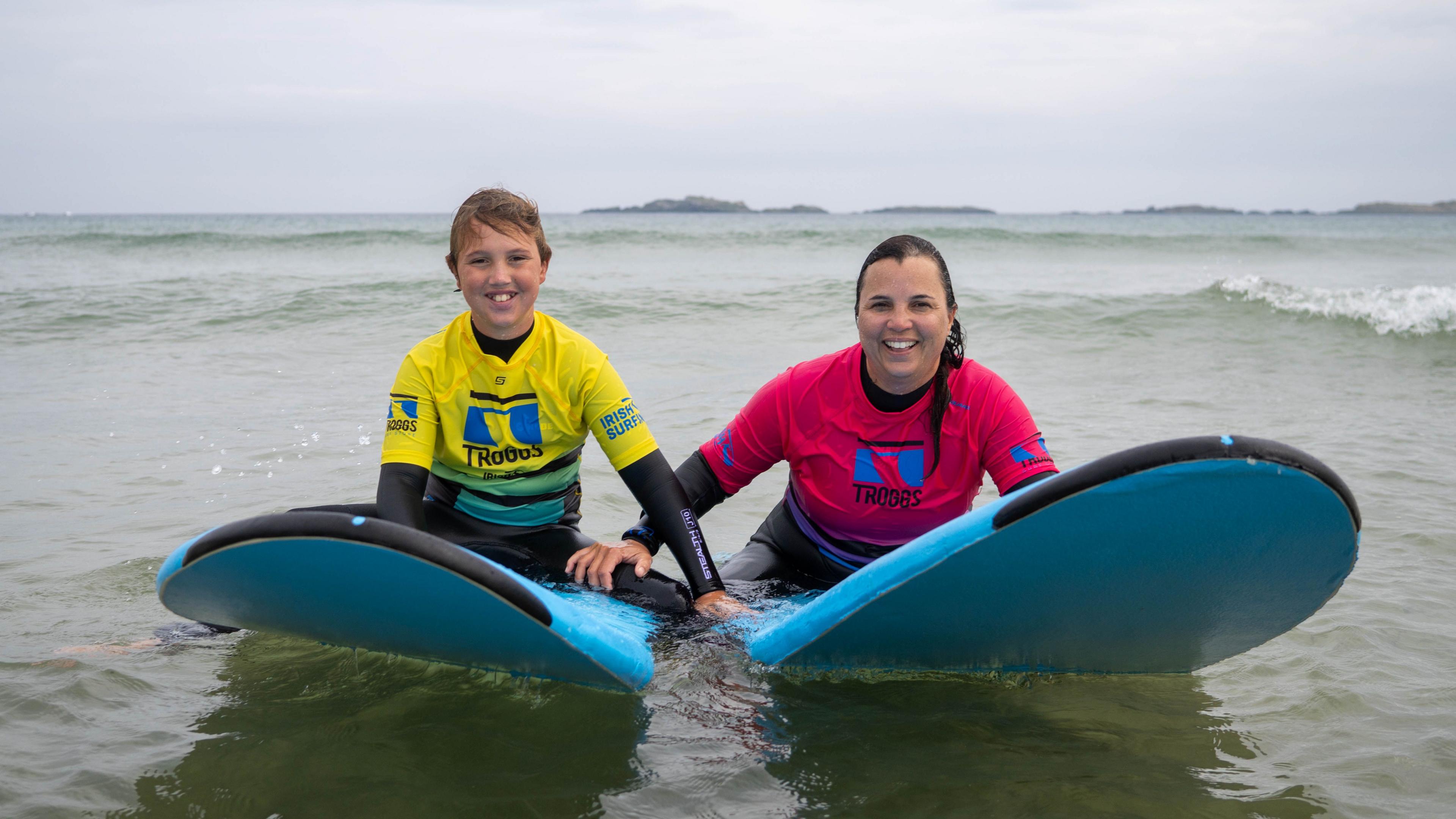 Boy smiling in a yellow wetsuit top on a blue board, beside his mum who is wearing a pink wetsuit top on her blue board