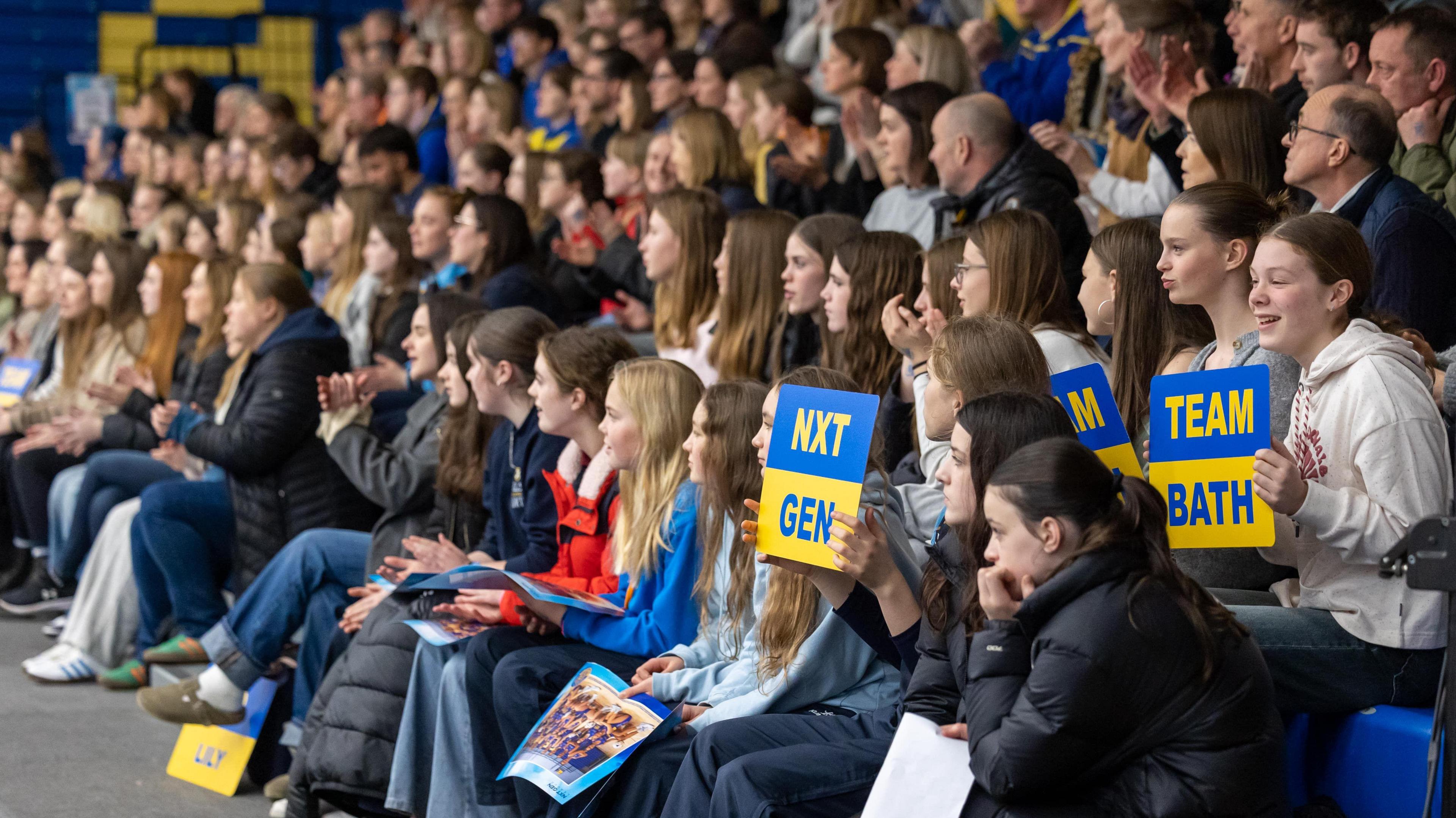 A large crowd of netball fans, most of them female, are seen packed into terraced seating as they watch Team Bath take on Nottingham Forest