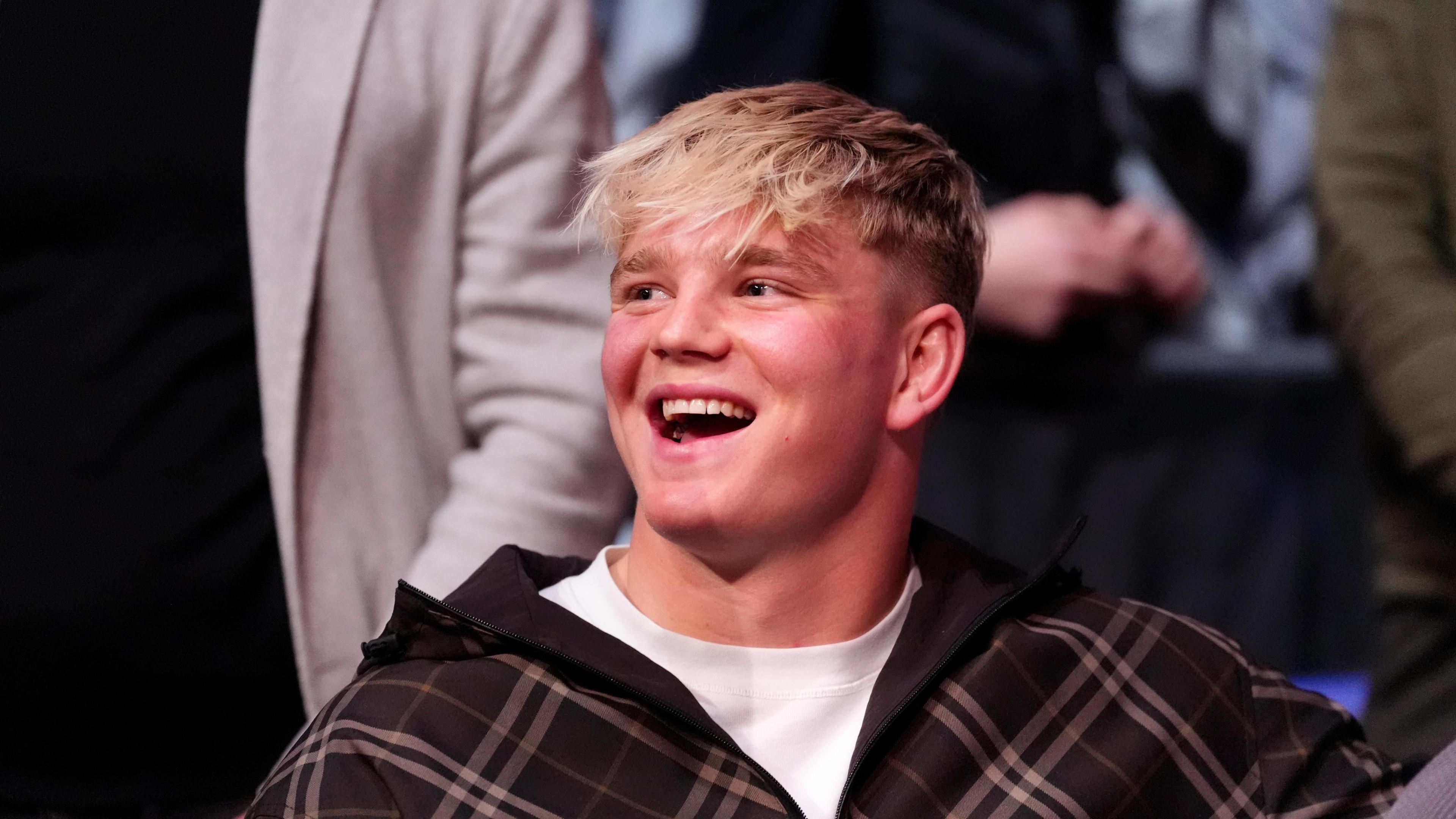 A young man smiling and looking away from the camera. He is wearing a white T-shirt and checked dark jacket. He has blonde hair. 