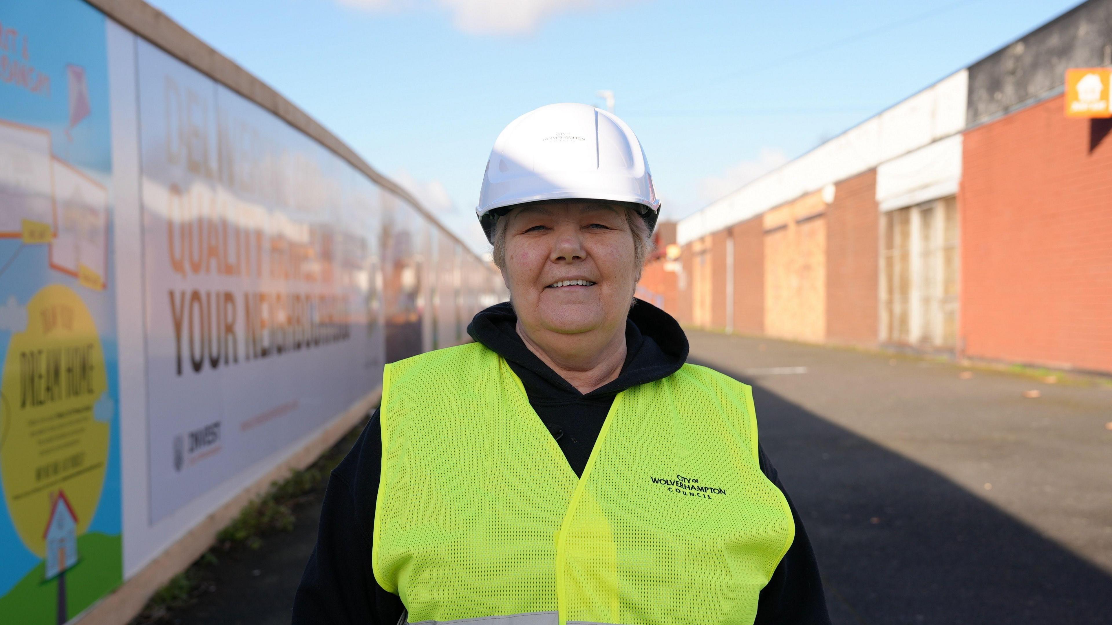 A woman wearing a yellow high-vis vest and white hard hat. She is smiling into the camera and standing next to a fence that has advertisements on. 