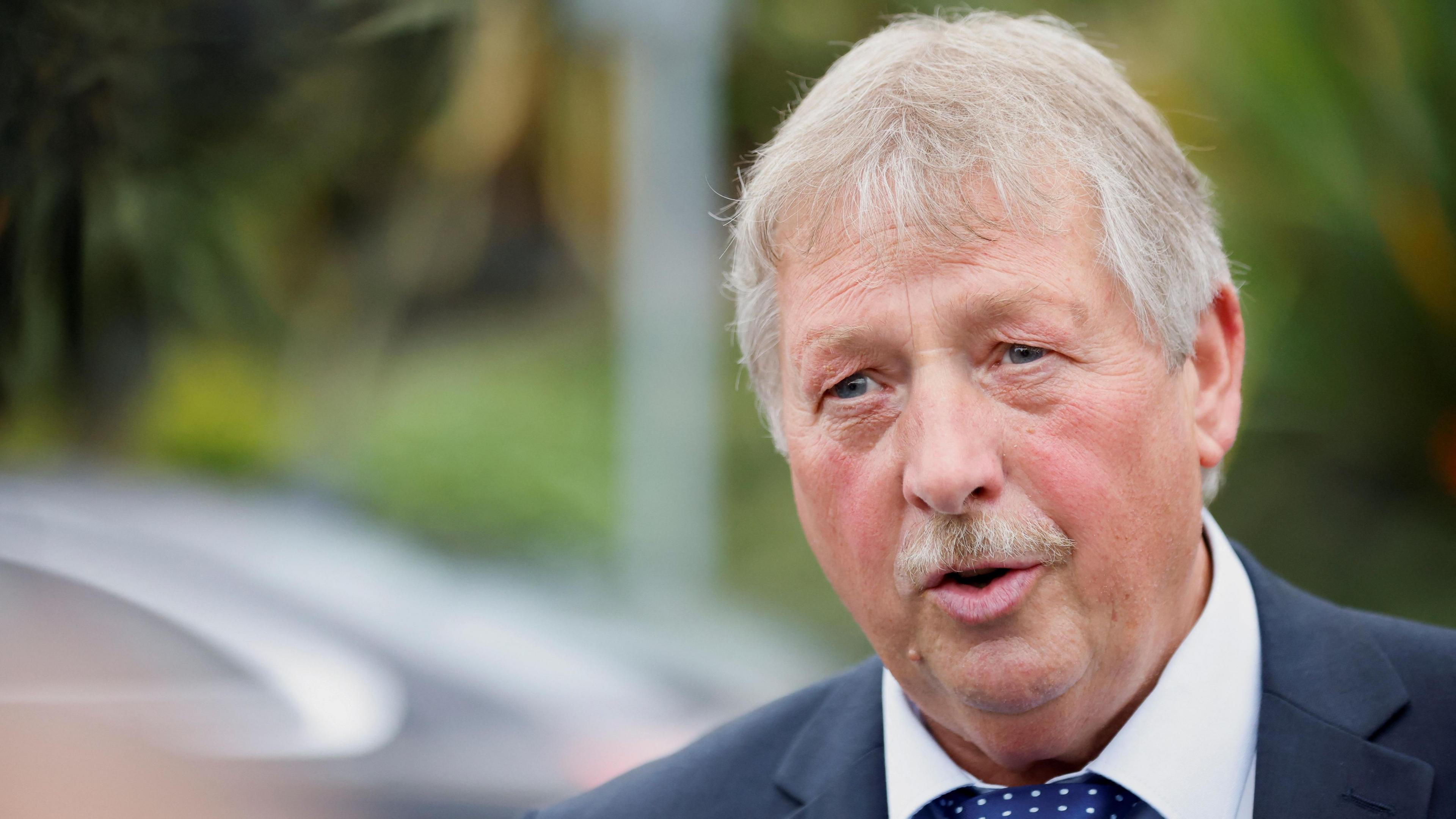 Sammy Wilson speaking. He is wearing a suit and tie. In the background a car and some trees are out of focus.