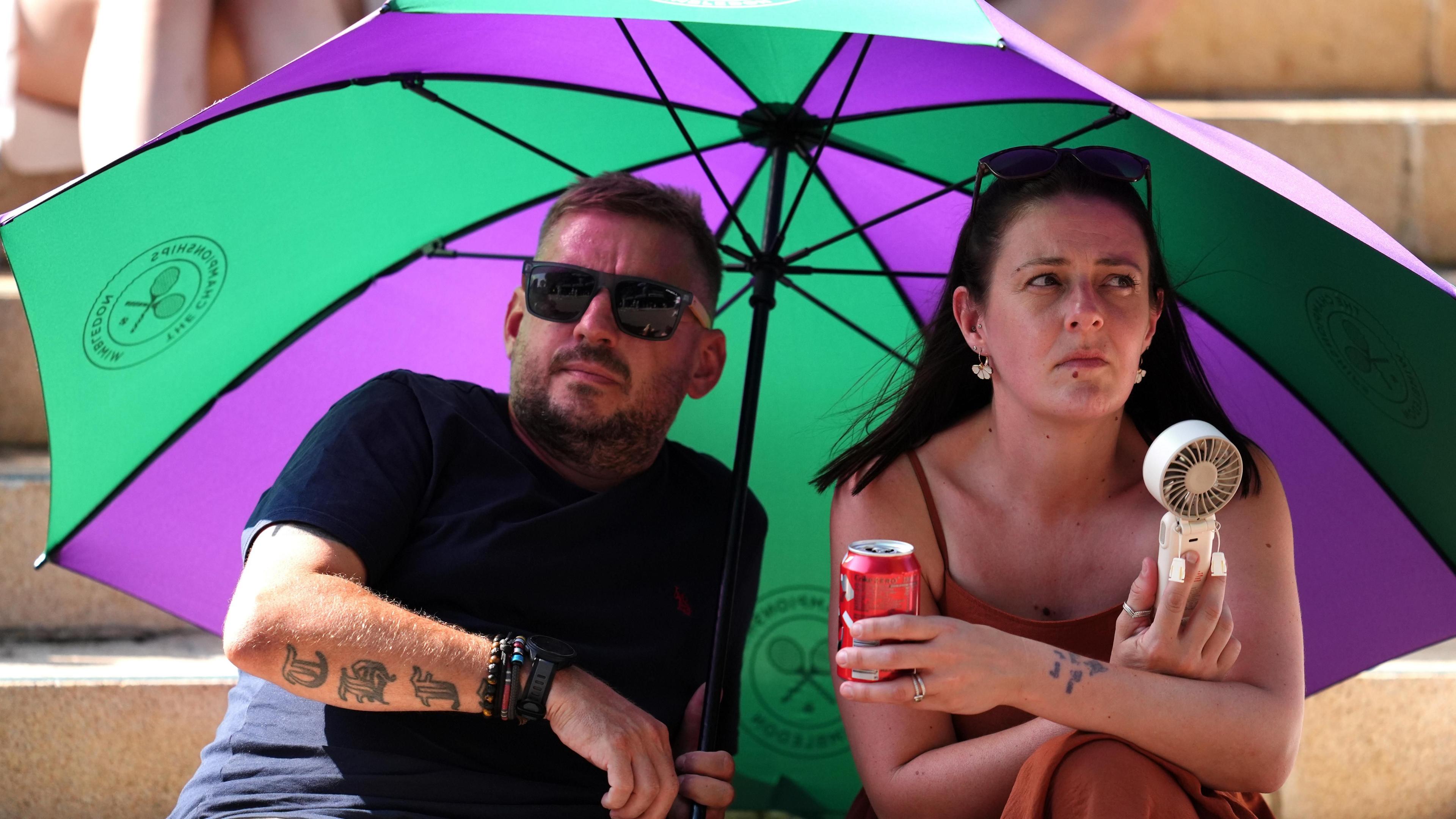 Spectators on The Hill at Wimbledon on Wednesday. A man and a woman are sat beneath the shade of a green and purple Wimbledon umbrella. The woman uses a hand-held fan. The man wears dark sunglasses.