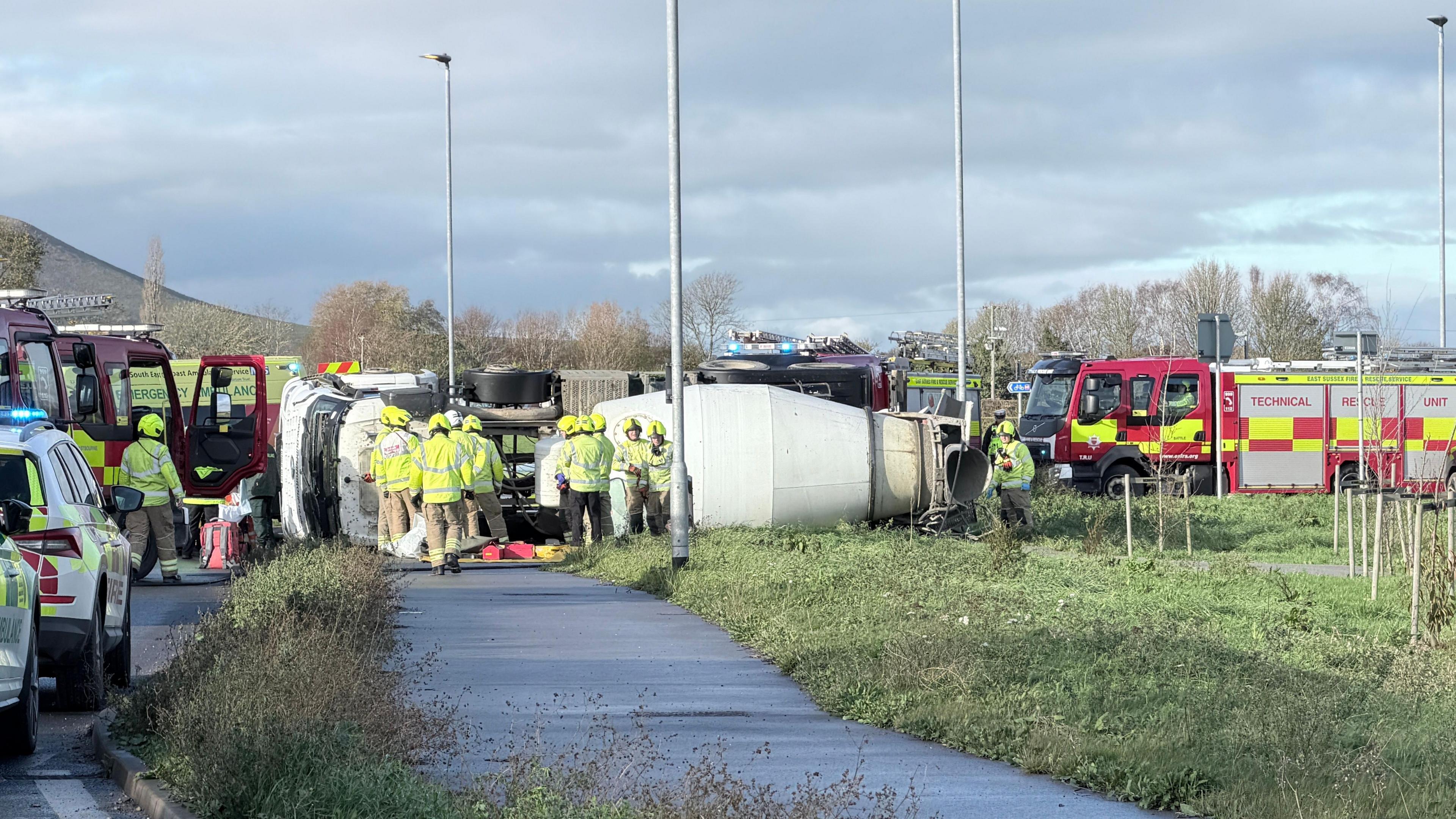 Emergency services vehicles gathered at the side of a road incident, with the overturned vehicle visible in the background. There is a fire engine on the right-hand-side as well as police cars on the left-hand-side. Fire crew can be seen surrounding the vehicle.