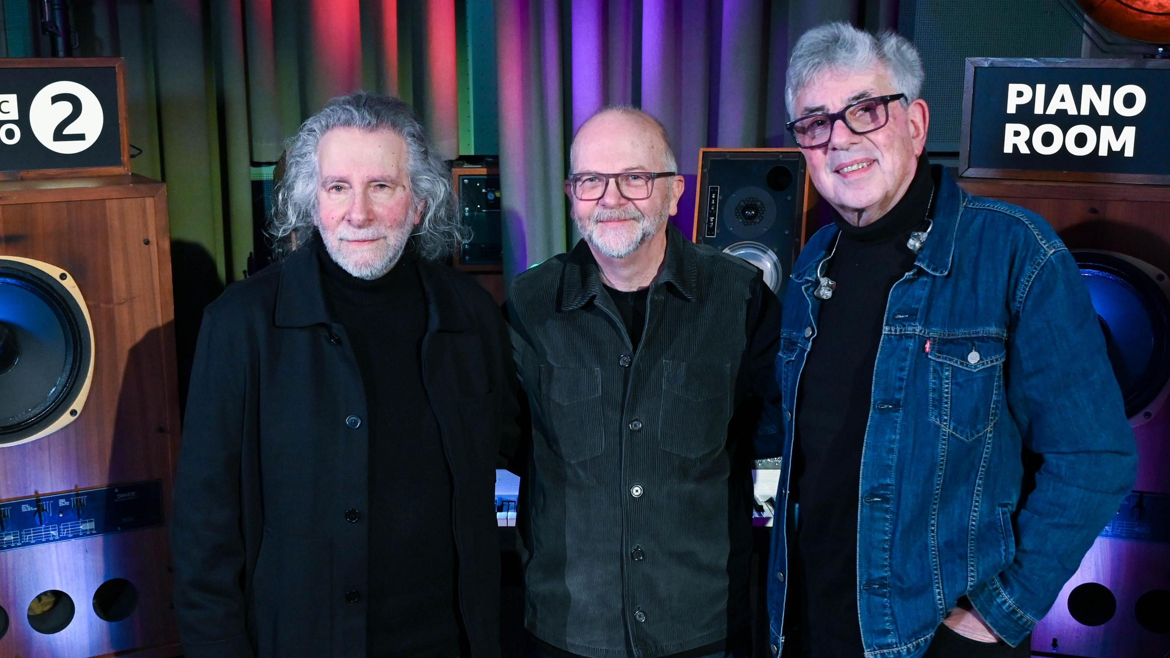Three members of 10cc smile for the cameras. They are standing in front of signage for BBC Radio 2 and Piano Room