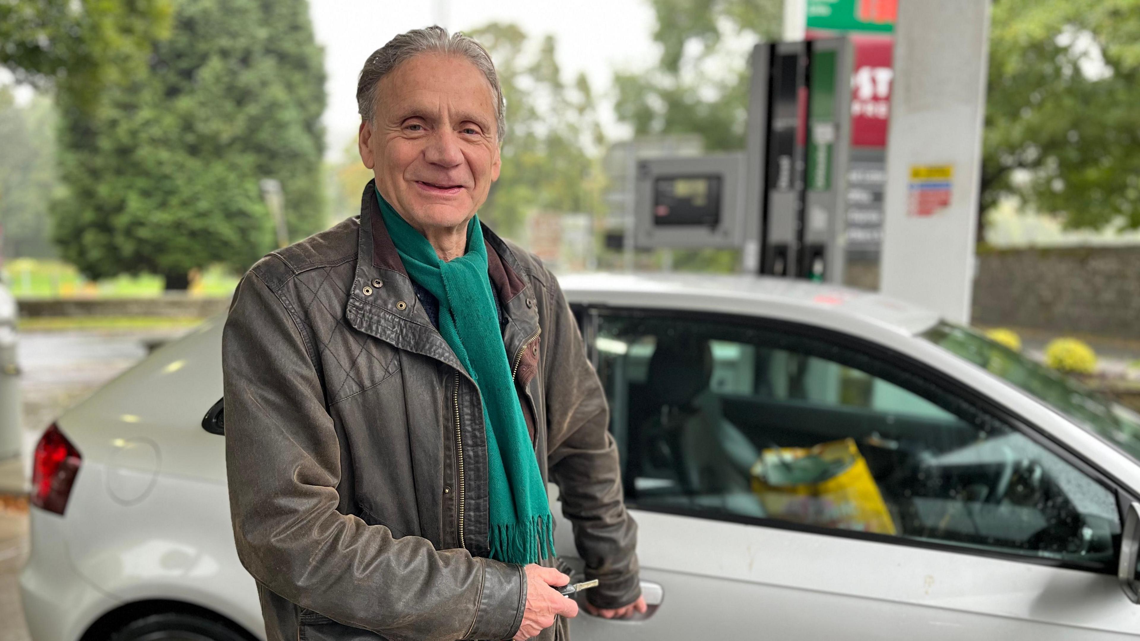 A man wearing a brown leather jacket and green scarf stands next to his silver car on a petrol station forecourt with fuel pumps in the background.