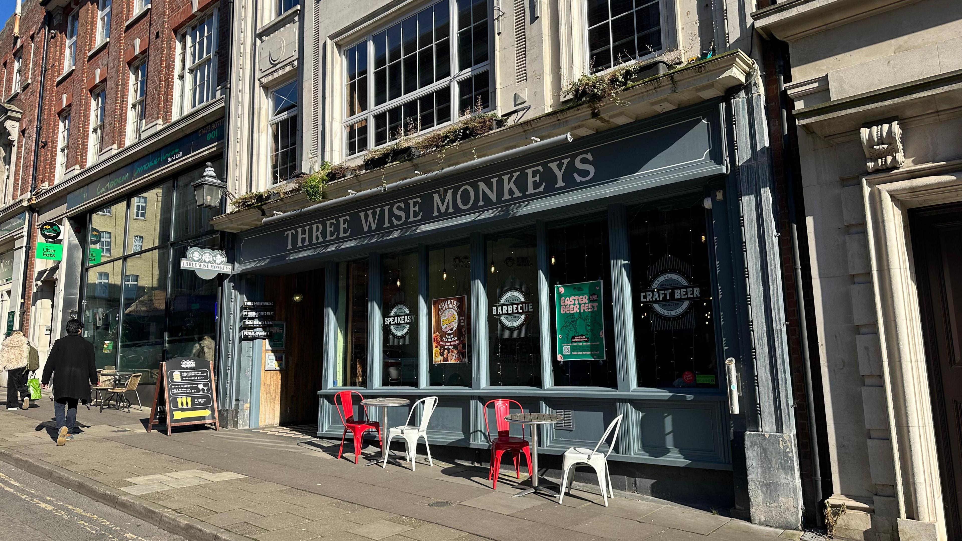 A general view of the outside of Three Wise Monkeys in Ipswich. Chairs and tables sit outside the pub which has large windows. Signage has been placed on the inside the window.