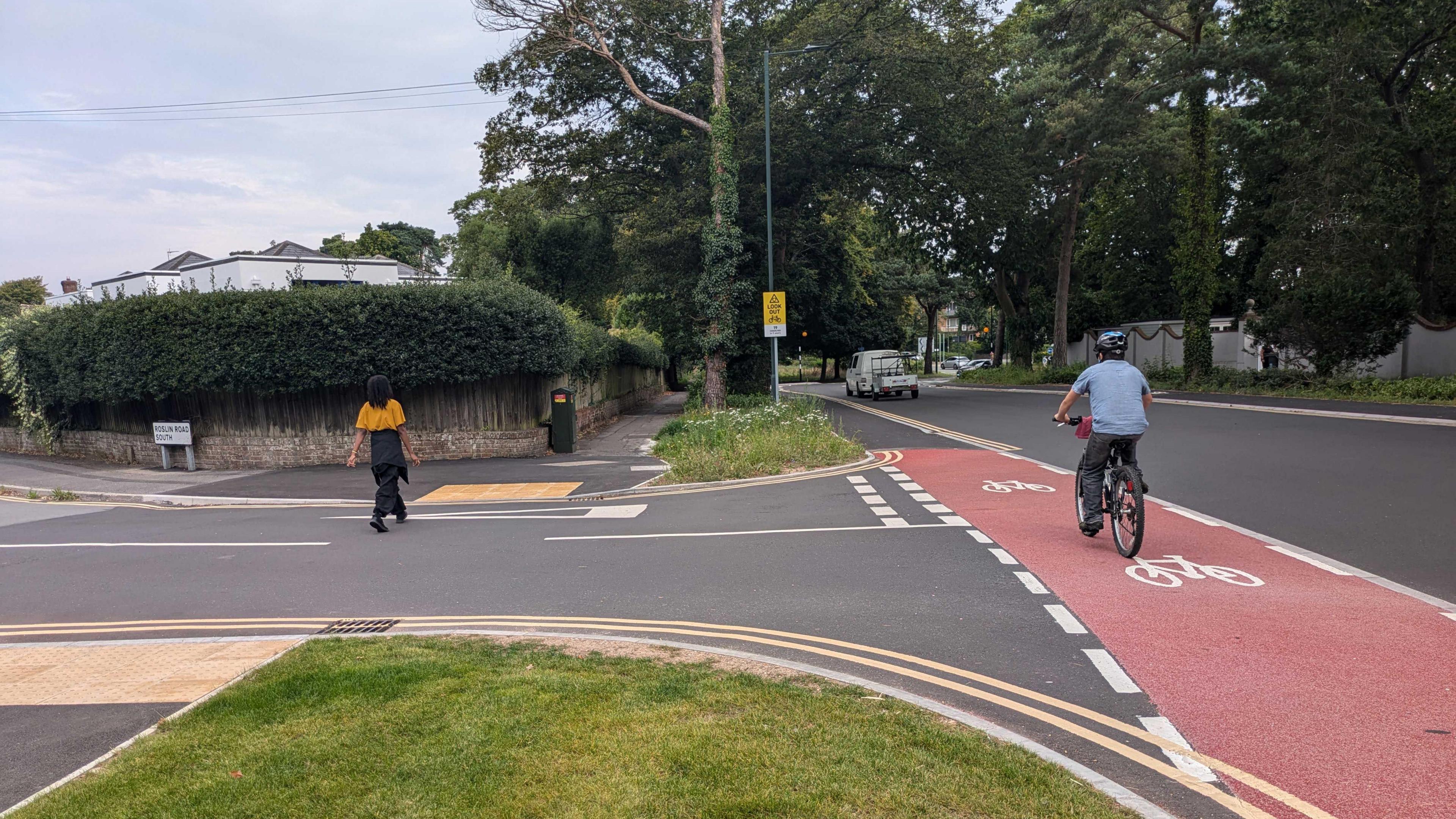 Improved junction and cycle lane at Glenferness Avenue and Roslin Road South. A man is riding a bike in the cycle lane. A woman is crossing the street. It's a cloudy day.