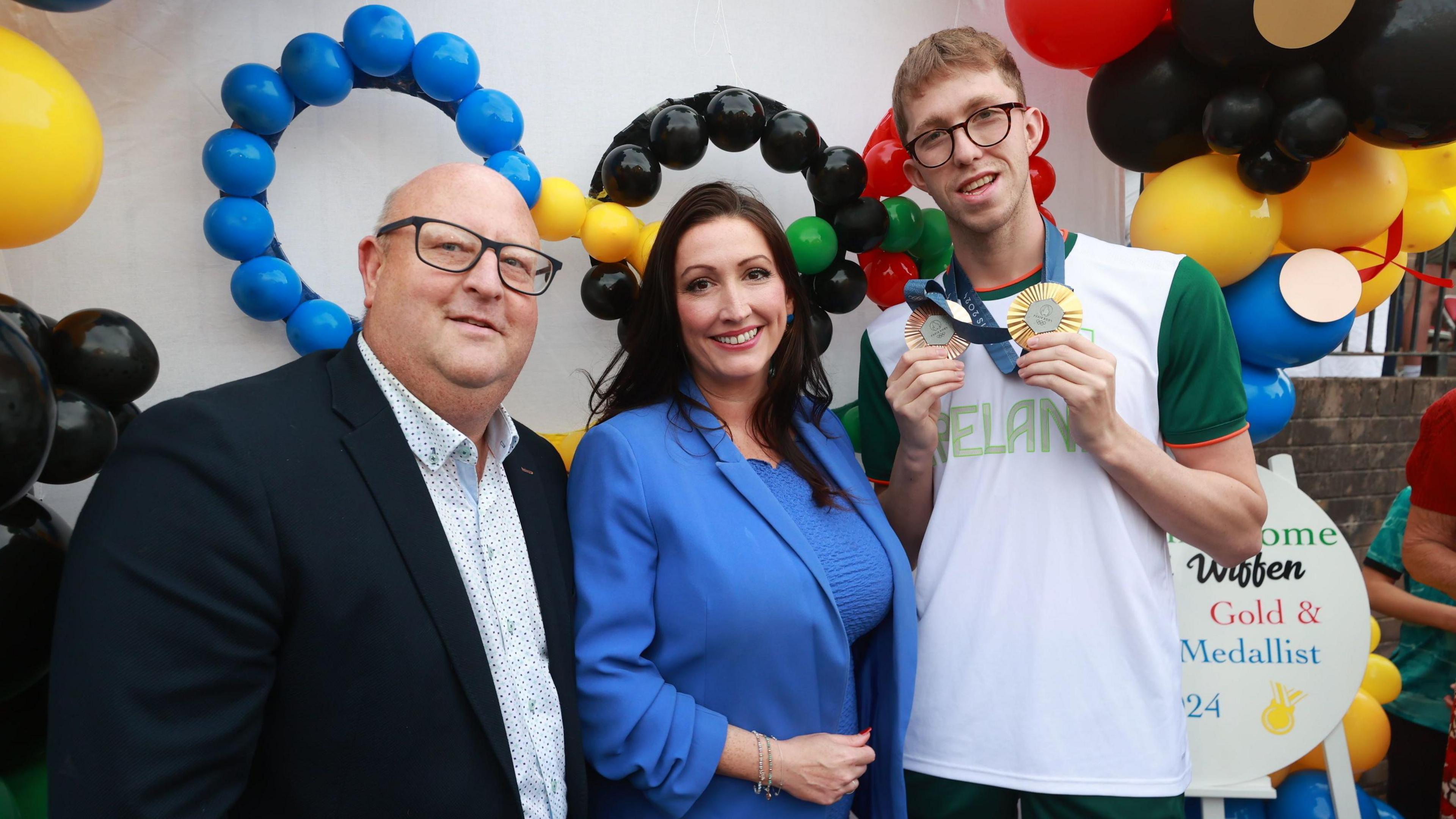 Daniel Wiffen stands beside the Deputy First Minister Emma Little-Pengelly (middle) and the DUP Councillor for Lurgan Peter Haire (left). They are all posing in front of a balloon arch and Daniel is holding up his medals.