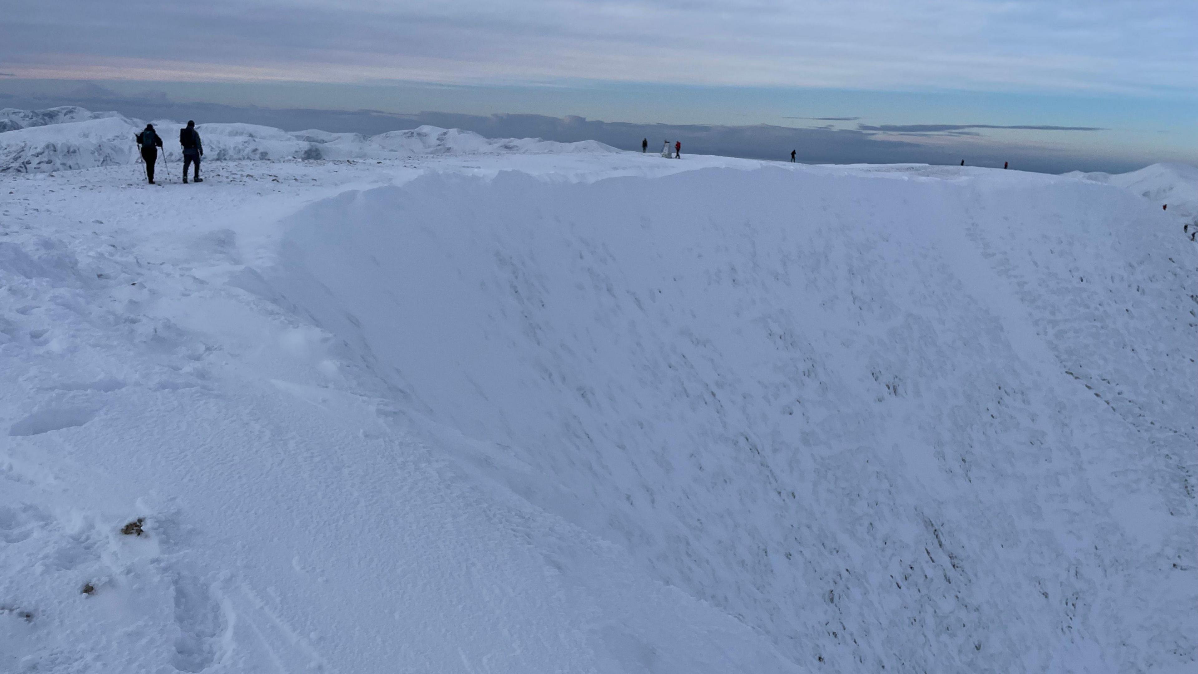 Helvellyn summit in full winter conditions. Snow covers the full summit as two walkers walk into the distance.