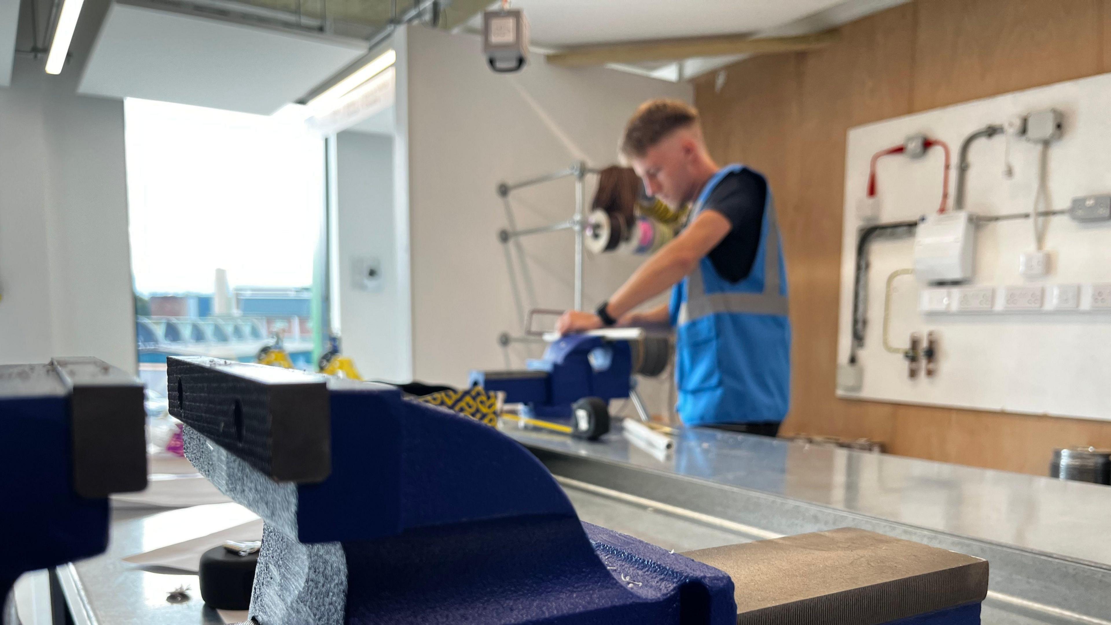 Man with hi-visibility top working at a vice with another vice in the foreground in a college workshop