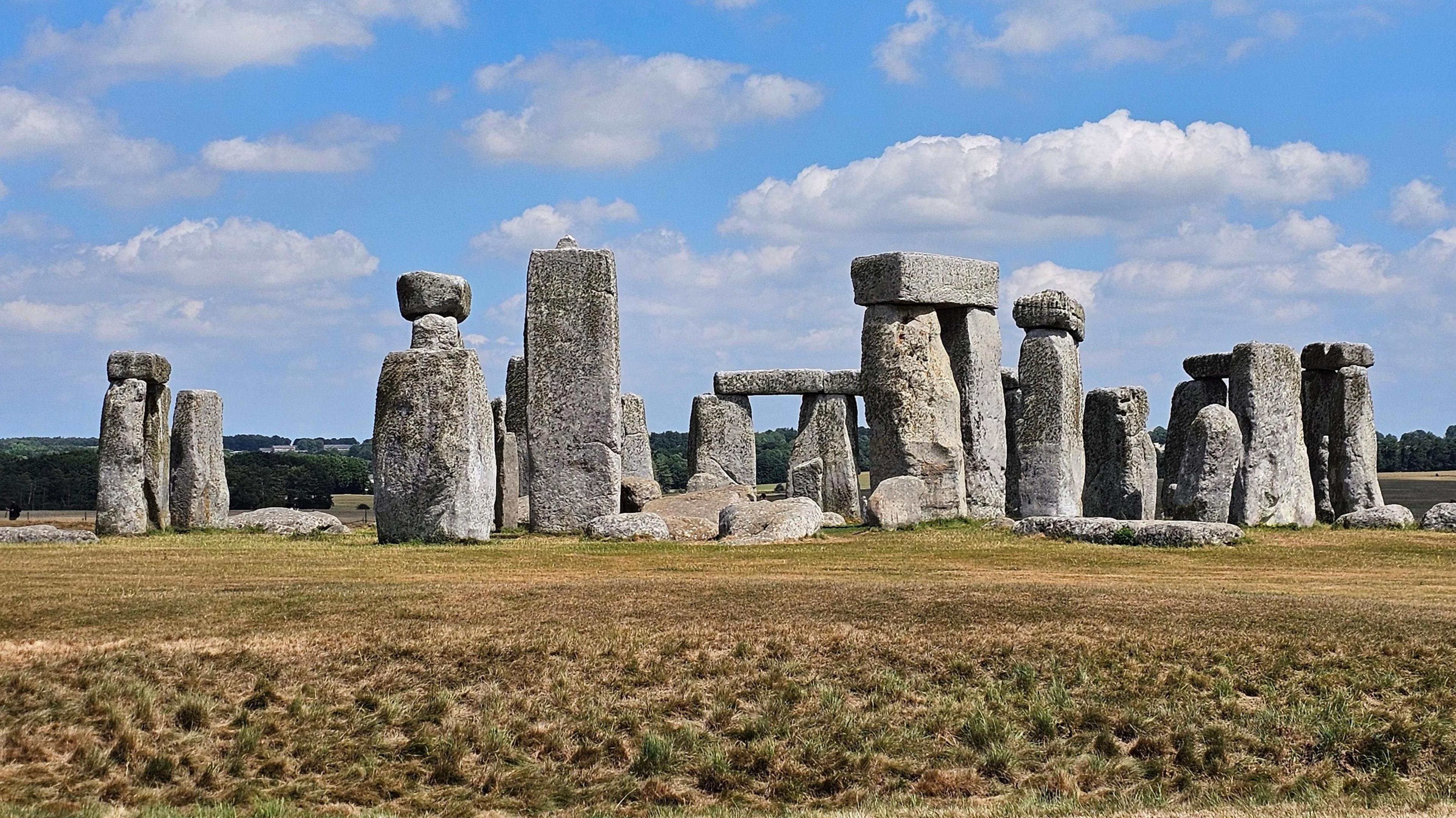 The stones at Stonehenge with yellowing grass in front. The sky is blue, with lots of white fluffy clouds.