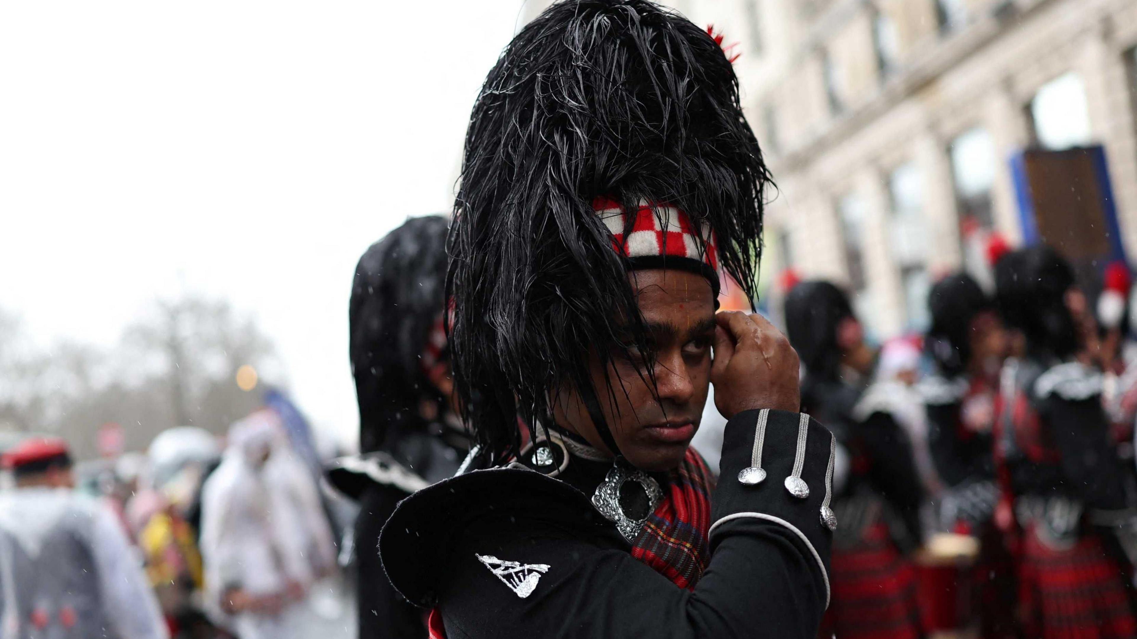 A man in traditional Highland attire with a very wet hat holds his hand to his face: in the background are members of his band in similar attire and people in rain ponchos. 