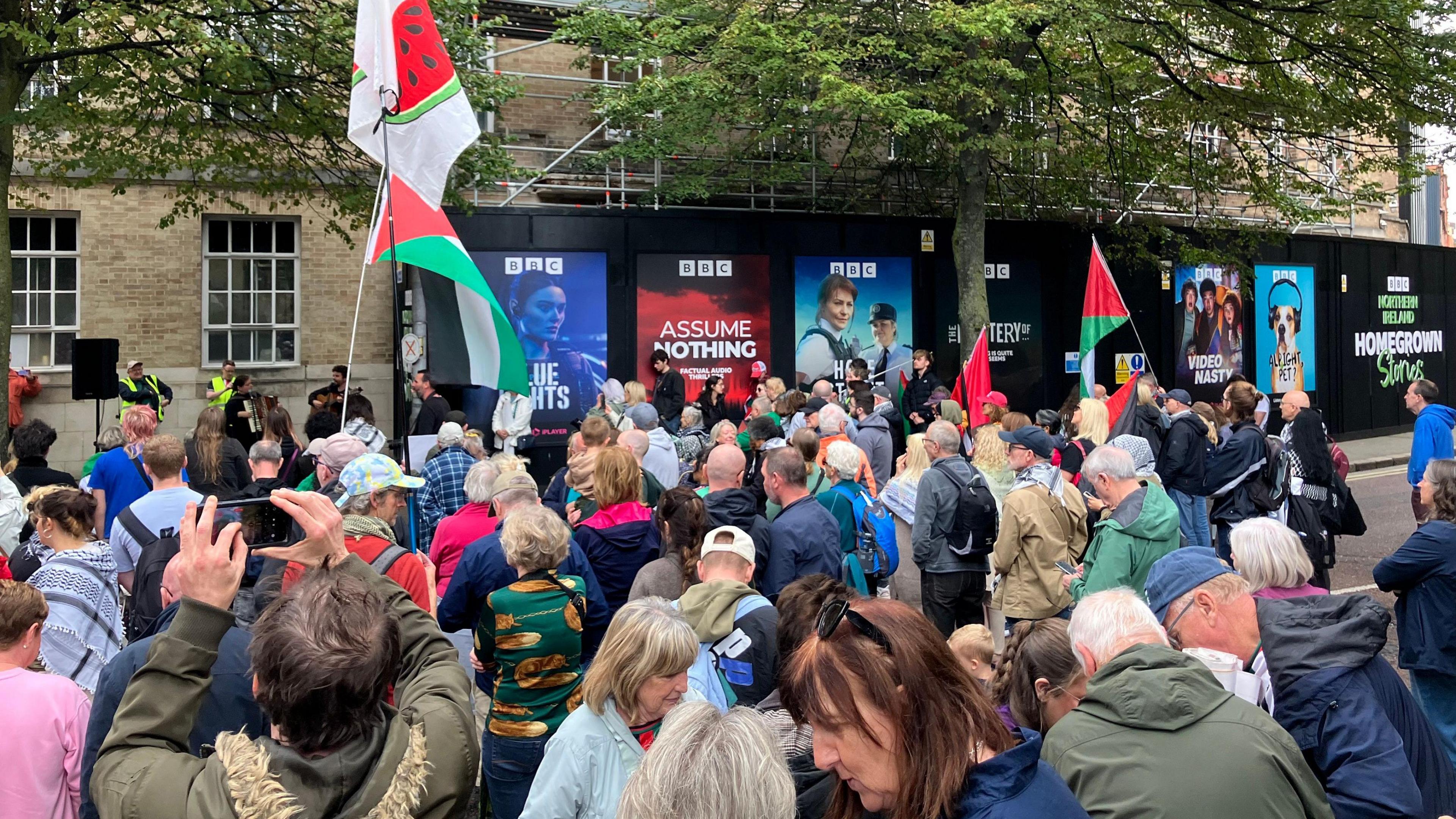 Crowds of protesters gathered outside BBC NI's headquarters in Belfast city centre. Some are holding Palestinian flags. There is scaffolding around the building.
