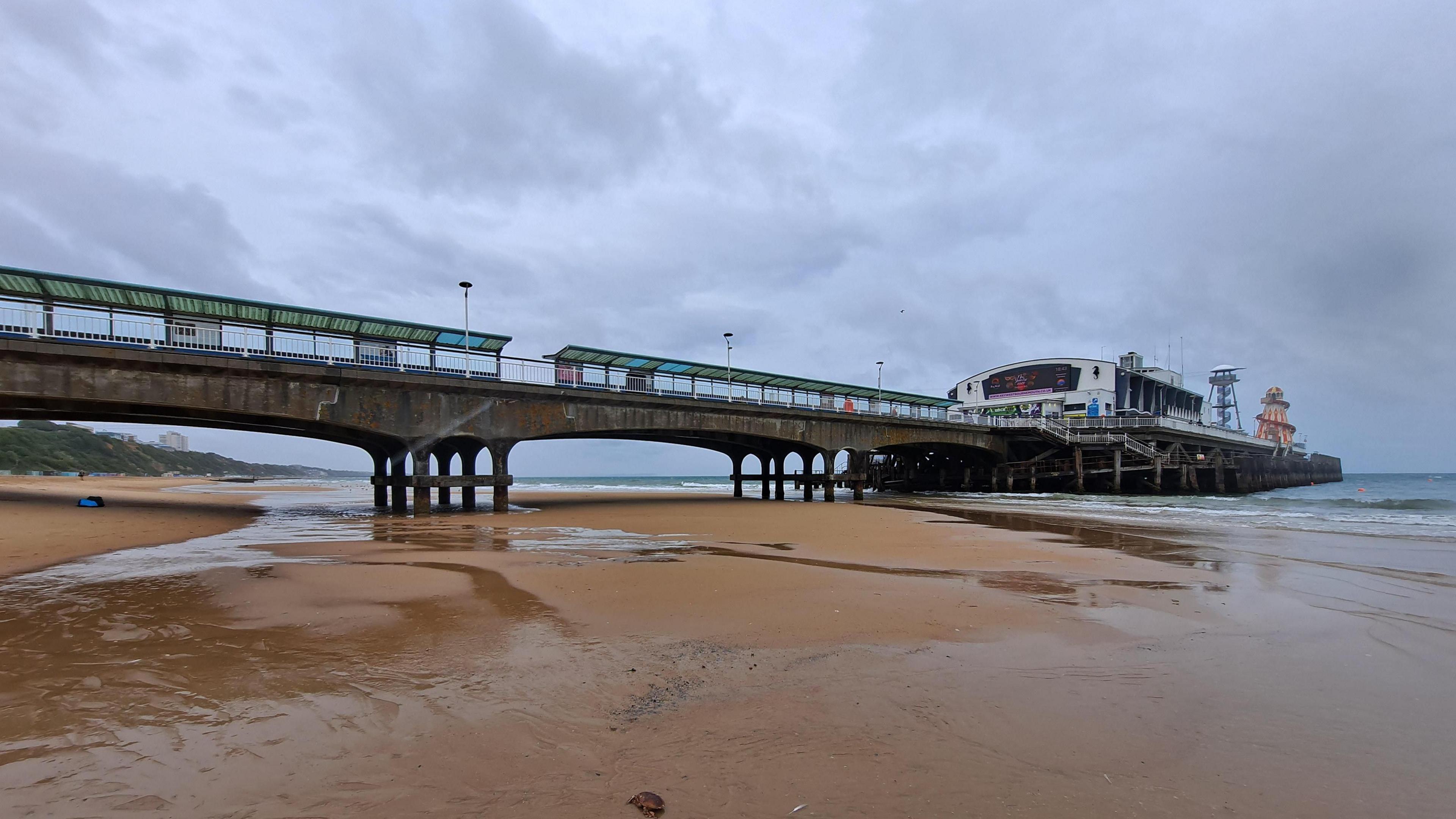 WEDNESDAY - Bournemouth Pier on a rainy day. The concrete pier sticks out into the sea at low tide. Overhead are grey clouds and it appears to have been raining. The wet sand on the beach and under the pier is empty. Under the end of the pier the sea is grey. At the end of the pier there is a theatre, a tower and a red and yellow striped helter-skelter. 