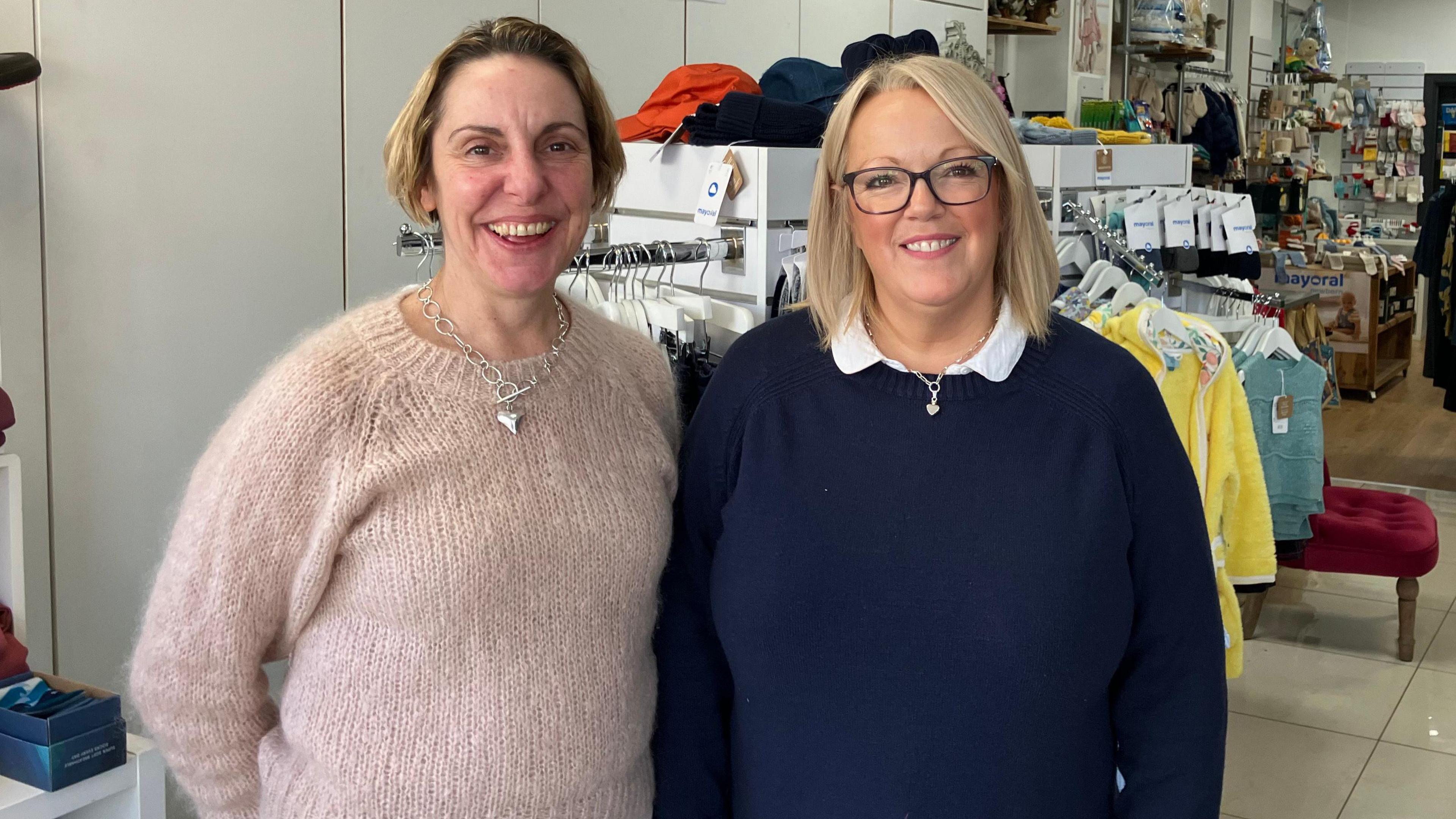 Photo of Emma Kemsley and Julie Burton who are independent traders in Lydney. They are smiling at the camera and standing shoulder to shoulder. One is wearing a light pink jumper and the other a dark blue jumper. Items for sale in a shop, including clothes and accessories, are visible in the background