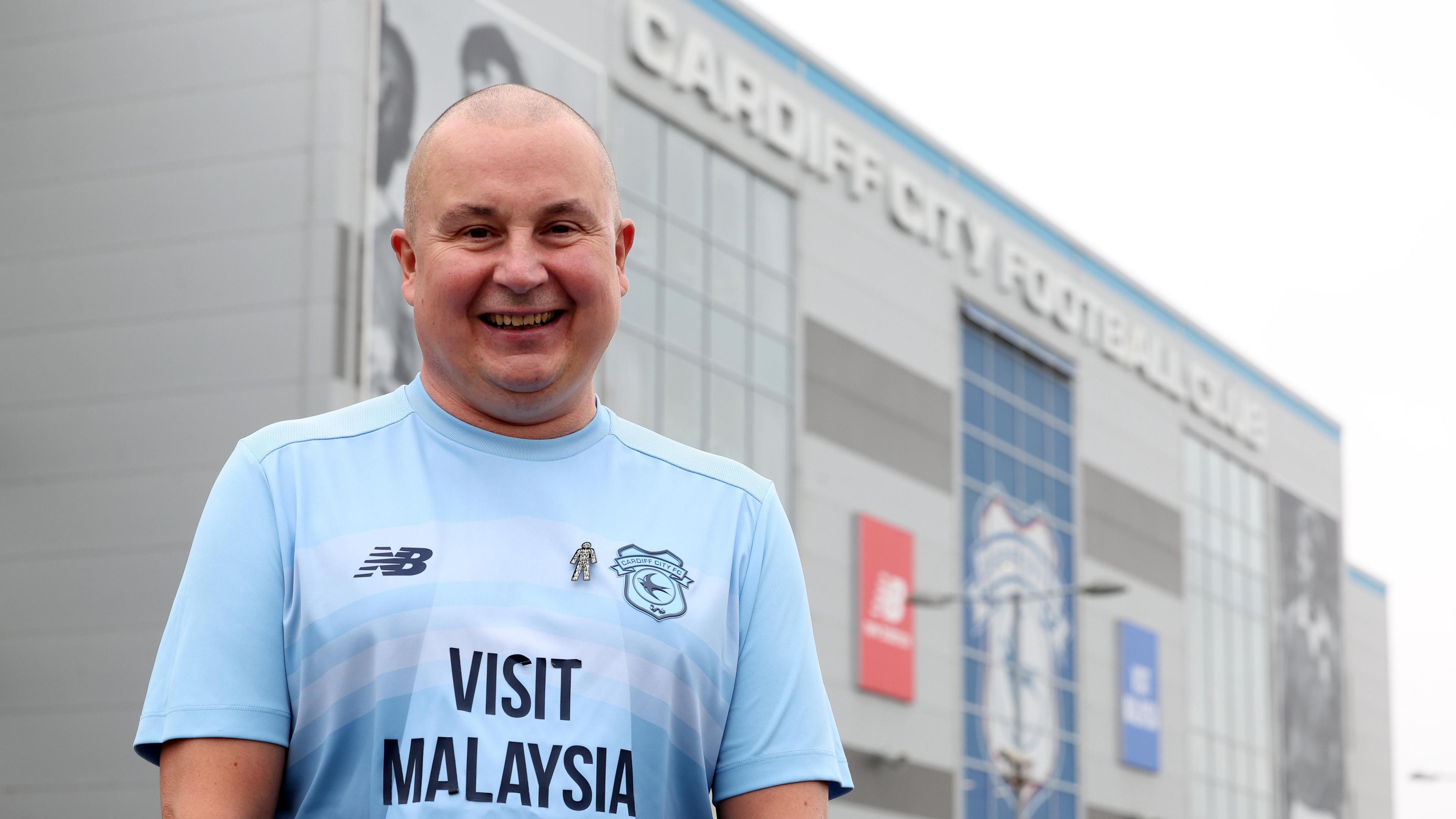 Mark Lewis is wearing a Cardiff City shirt as he stands outside the stadium. He is smiling.