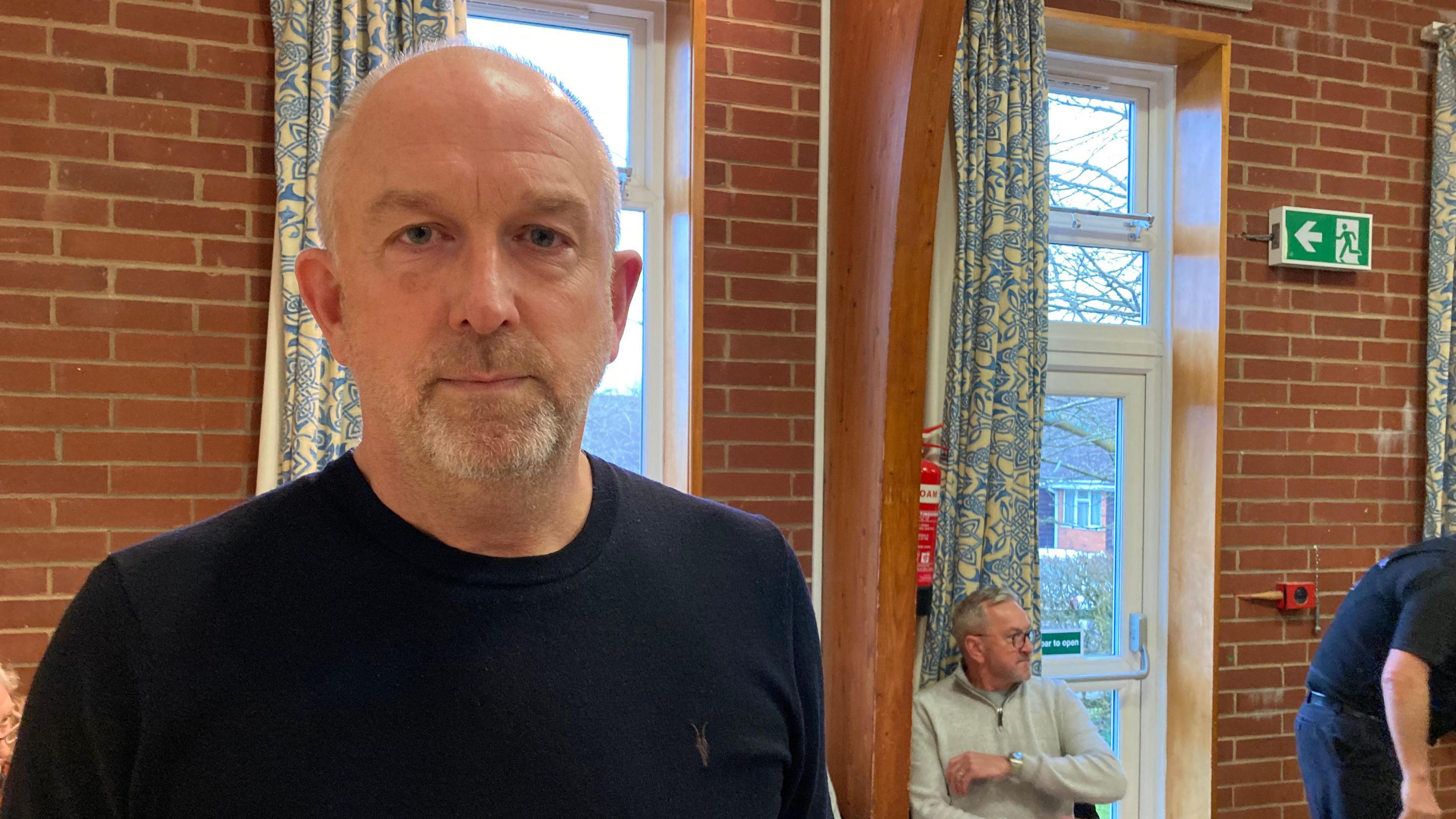 Steve is standing in a hall. He has very short grey hair with grey facial hair and is wearing a black sweater. He stands in front of glass windows and doors with a couple of men behind him in the background.