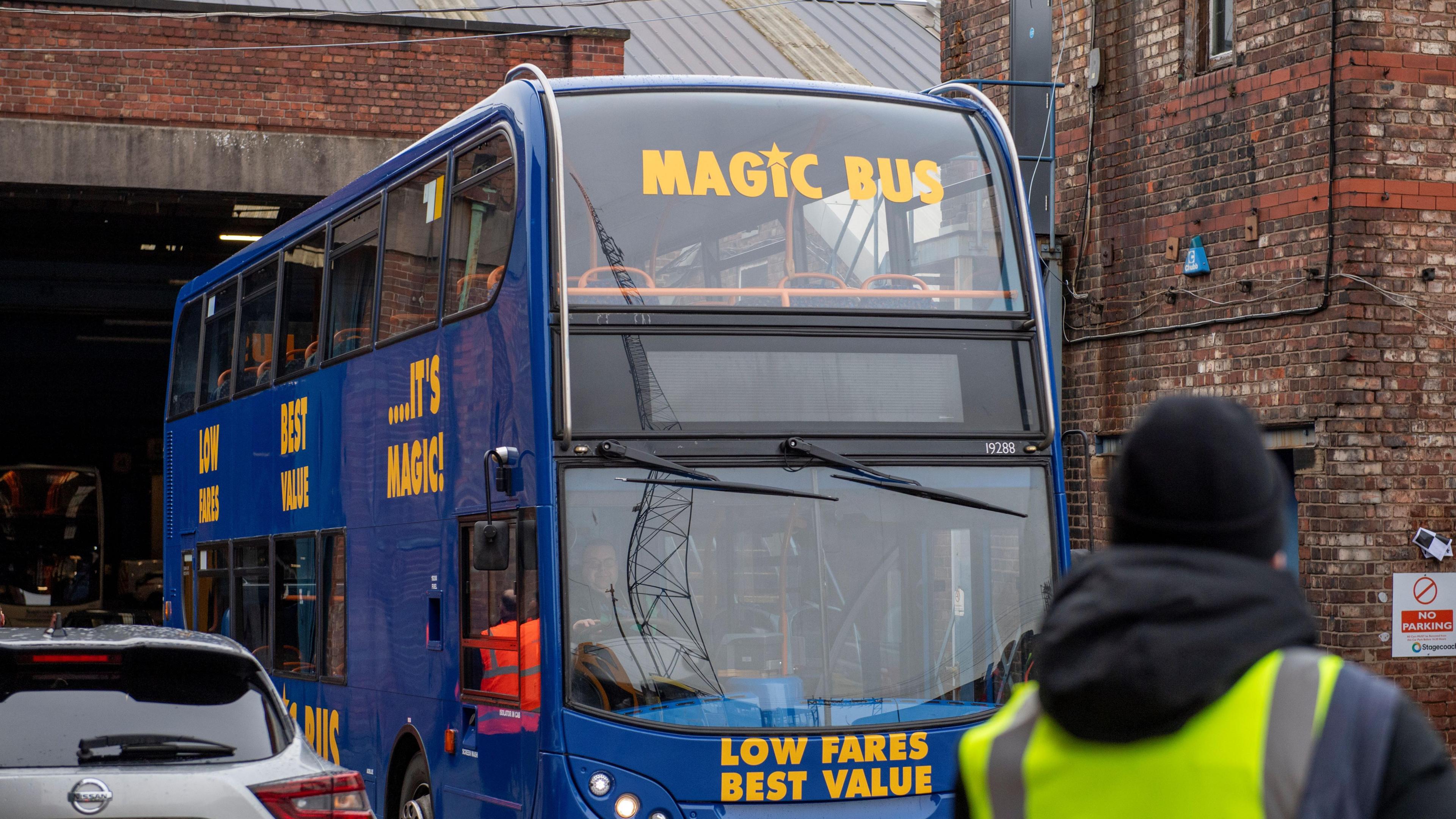 A man in a high-vis vest and black hat, looks on at the front of a blue-liveried Magic Bus. 