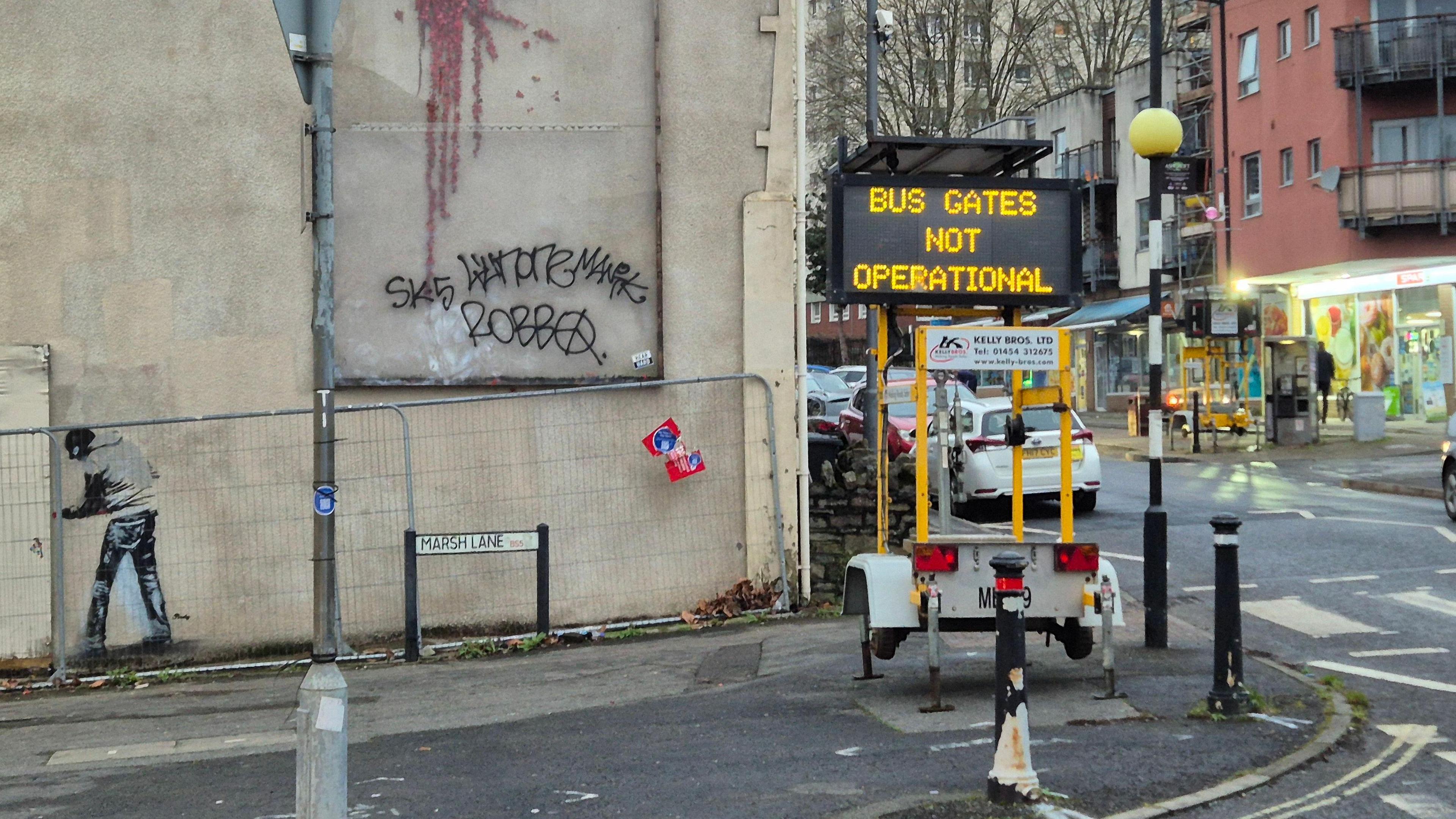 A temporary sign which reads 'bus gates not operational' stands on the pavement next to a wall, and sign for Marsh Lane.