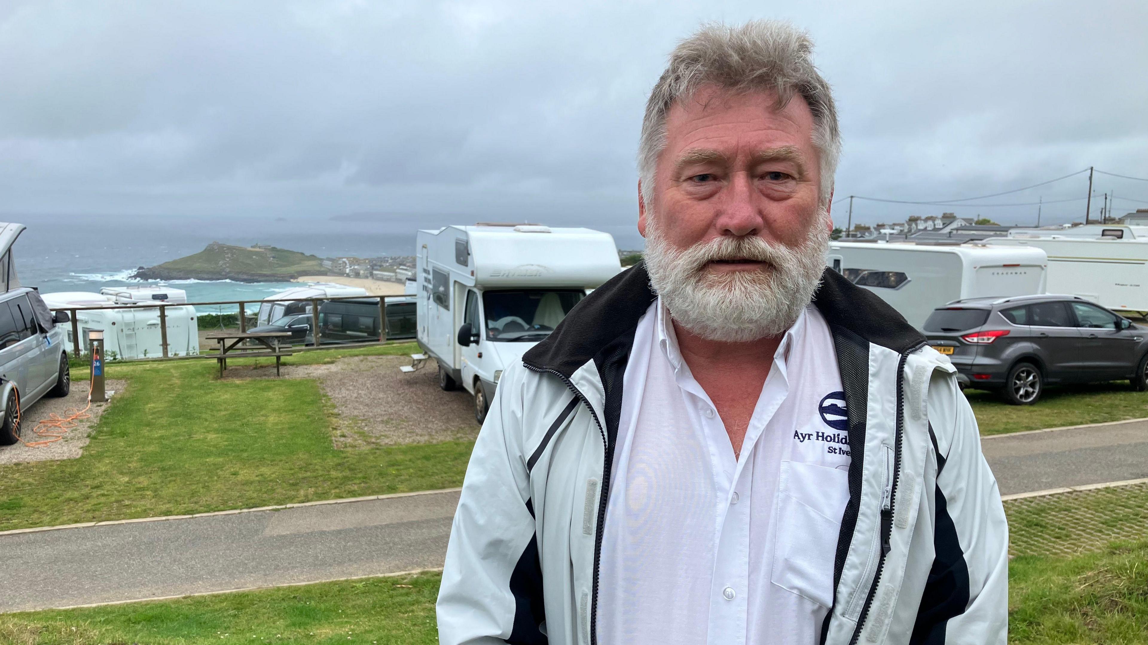 A man stands on a grassy areas with a coastal holiday park. The are motorhomes in the background and you can sea the ocean in the distance alongside the coastline.  There are grey clouds in the sky. 