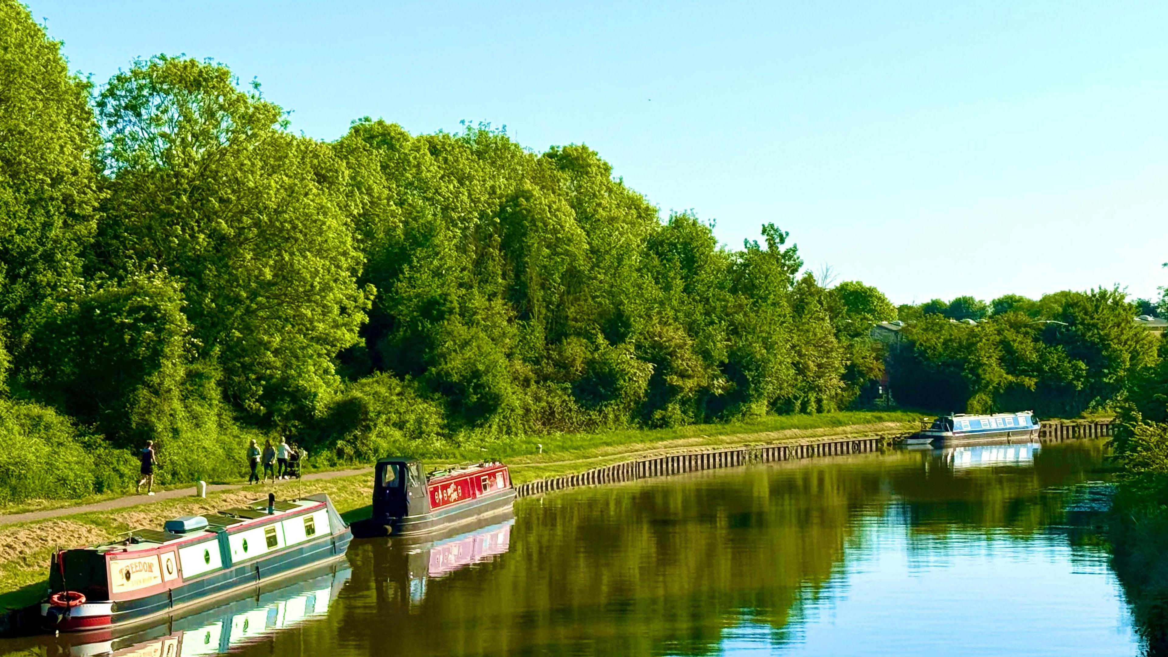 view of a canal with three canal boads with lots of green trees on the banks.  Blue skies and sunshine above