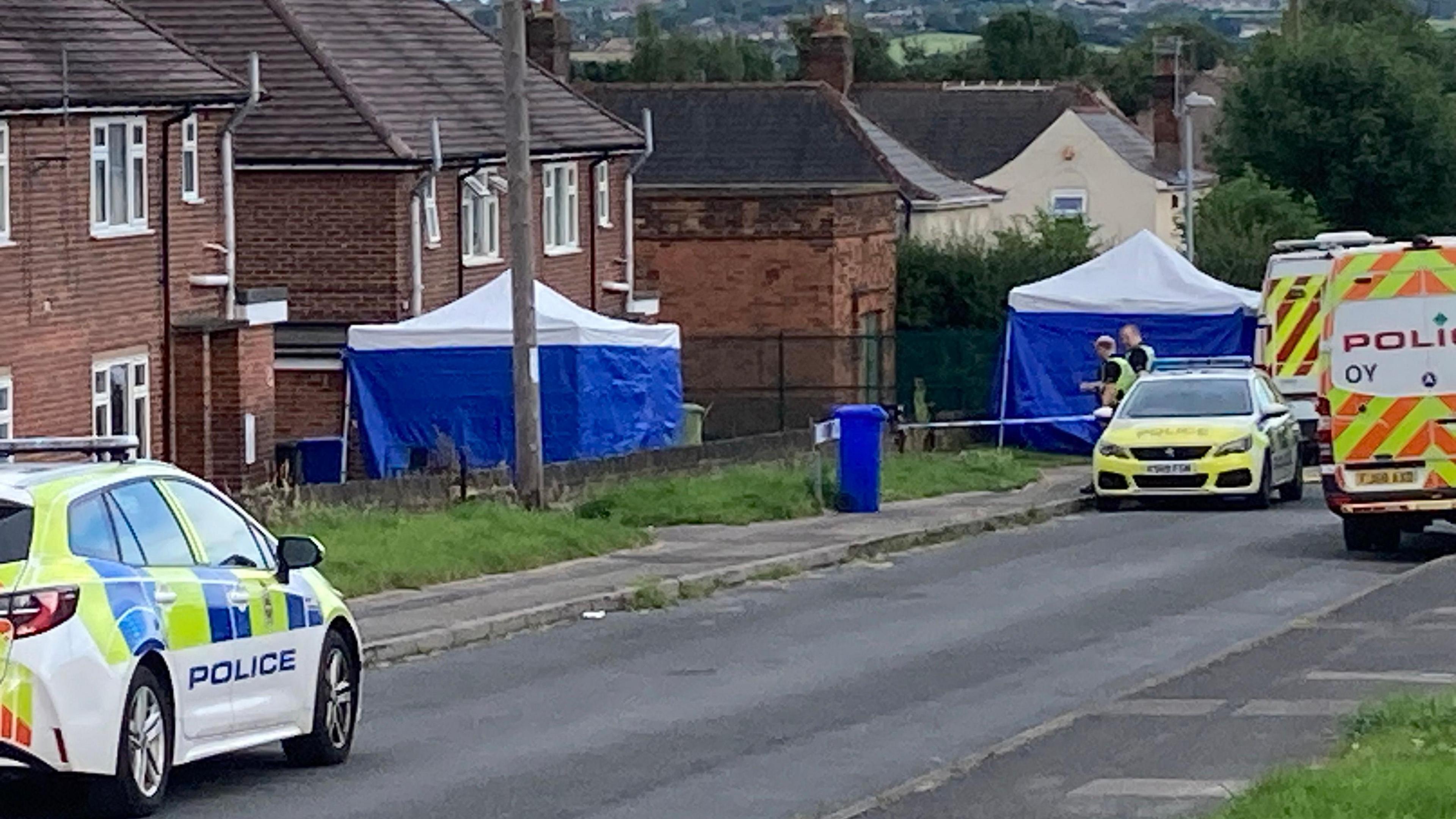 Two blue tents can be seen outside a house, with two police cars and two police vans parked on the street 