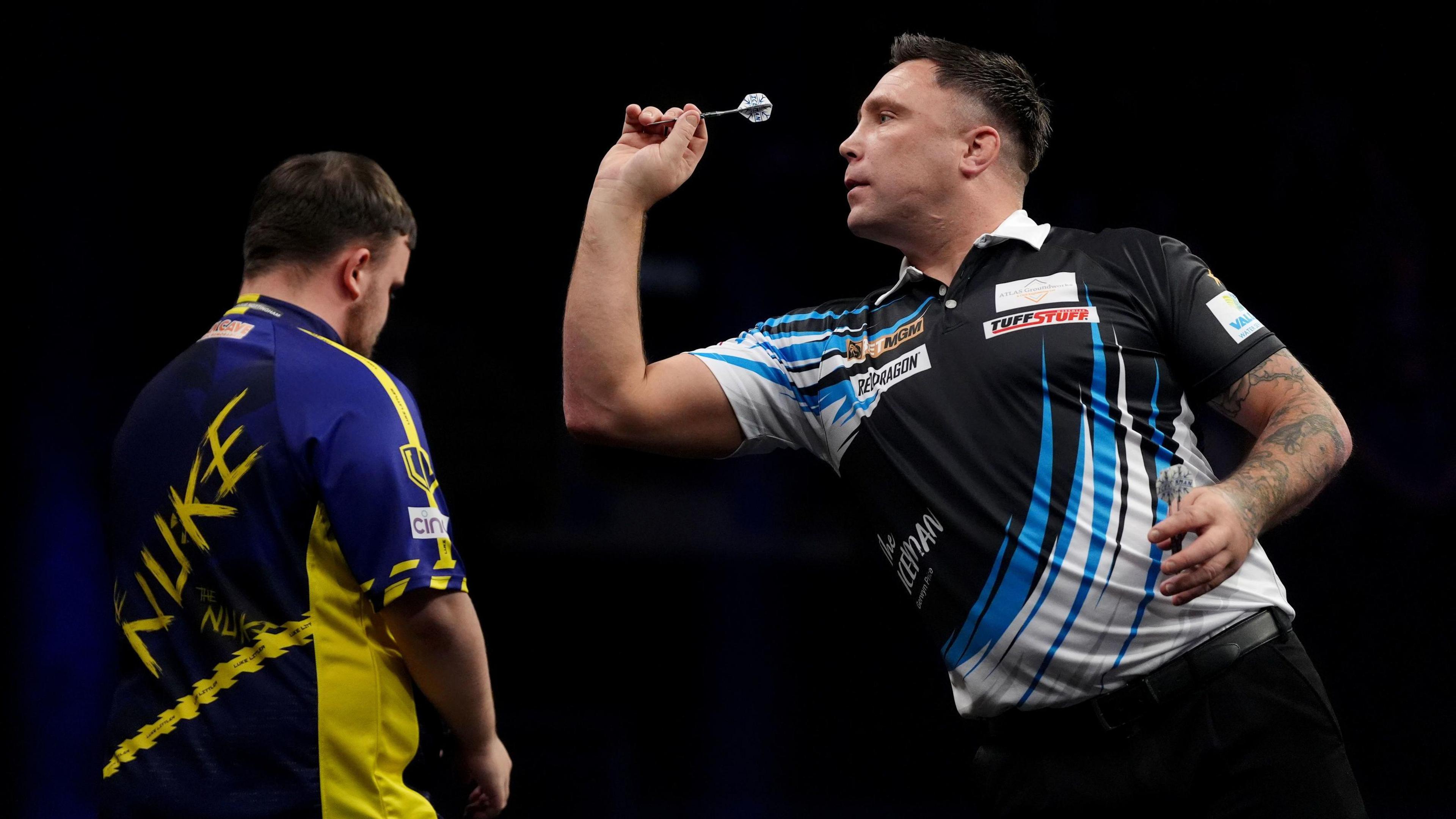 Gerwyn Price, pictured throwing a dart at a dartboard out of shot. He is wearing a black shirt with blue and white shards across it and a white collar. Luke Littler has his back to the camera, looking straight ahead. The background is black. 