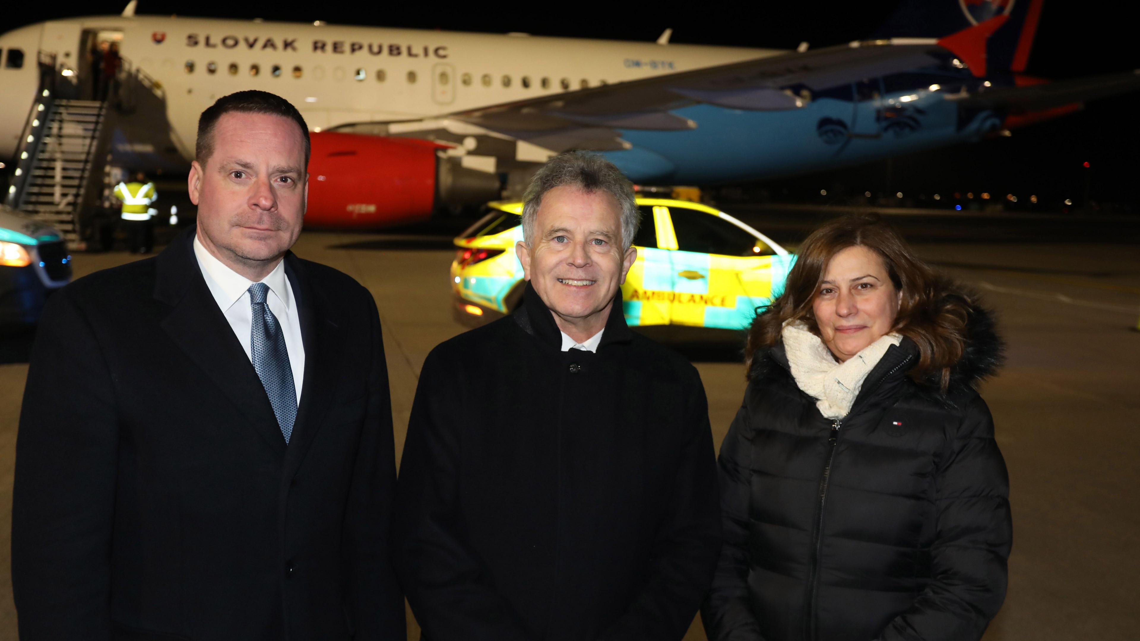 Two men and a woman standing on the tarmac at an airport in front of a place with the words "Slovak Republic" on the side. In front of the plane is an SUV with "ambulance" written on the site of it.