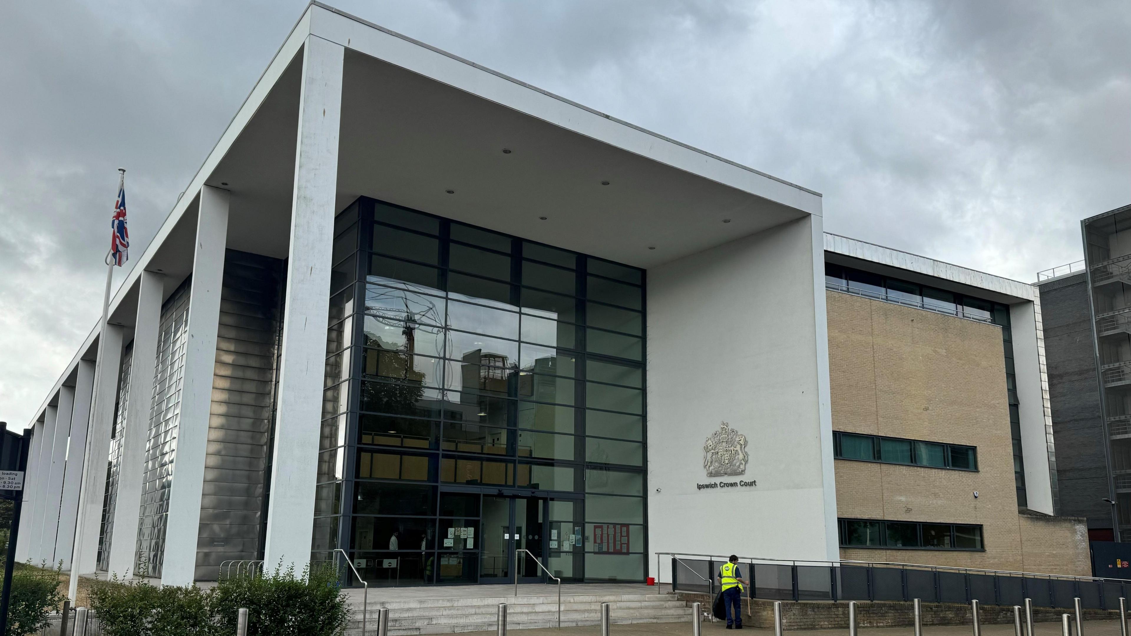 The front of Ipswich Crown Court, a modern building with a huge multi-storey glass facade. A person in a high-visibility jacket is standing outside with their back turned.