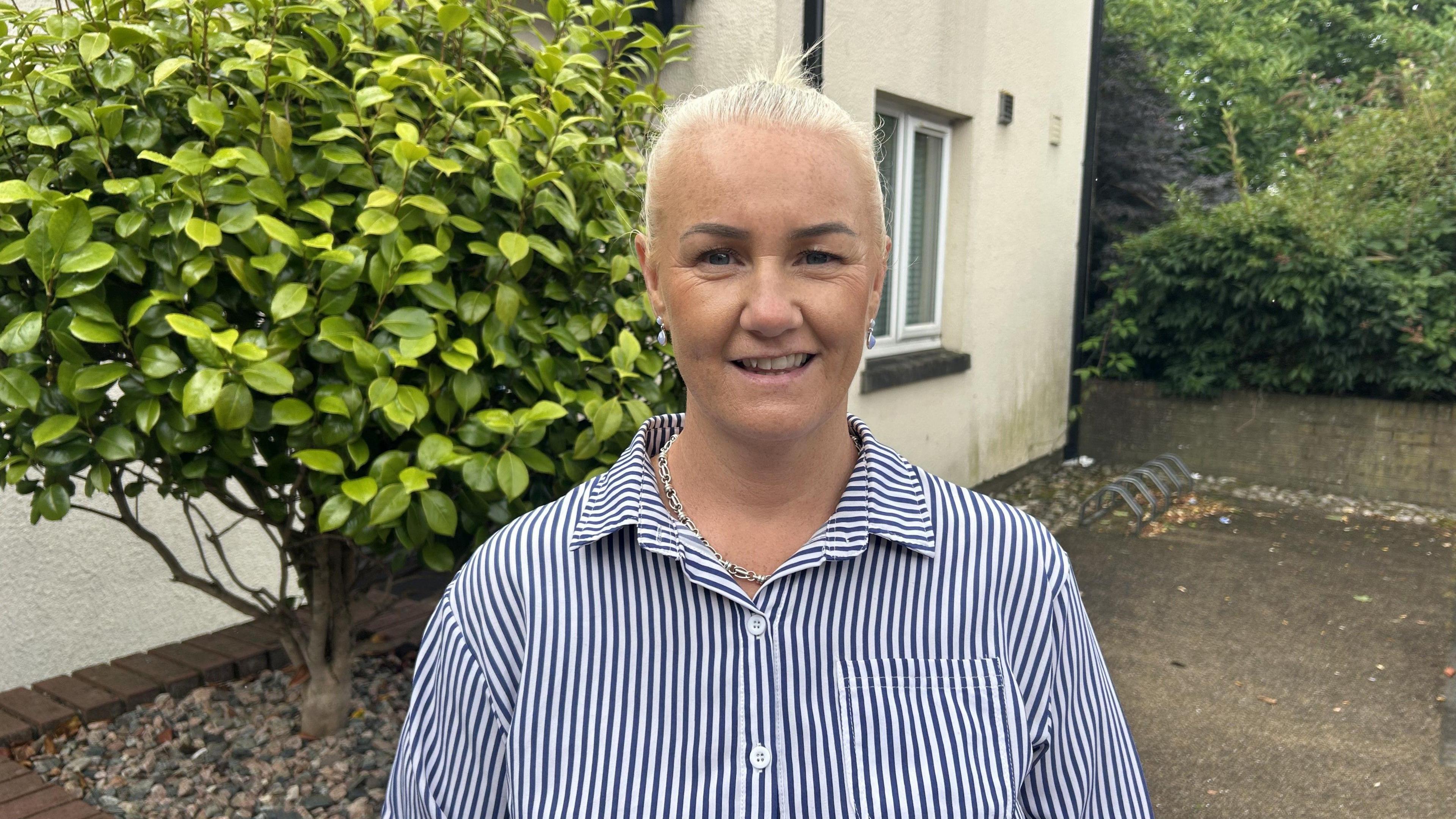 A woman with white hair smiles at the camera. She is wearing a navy and white stripy shirt. In the background you can see the side of a white house with a green tree the right of the photo