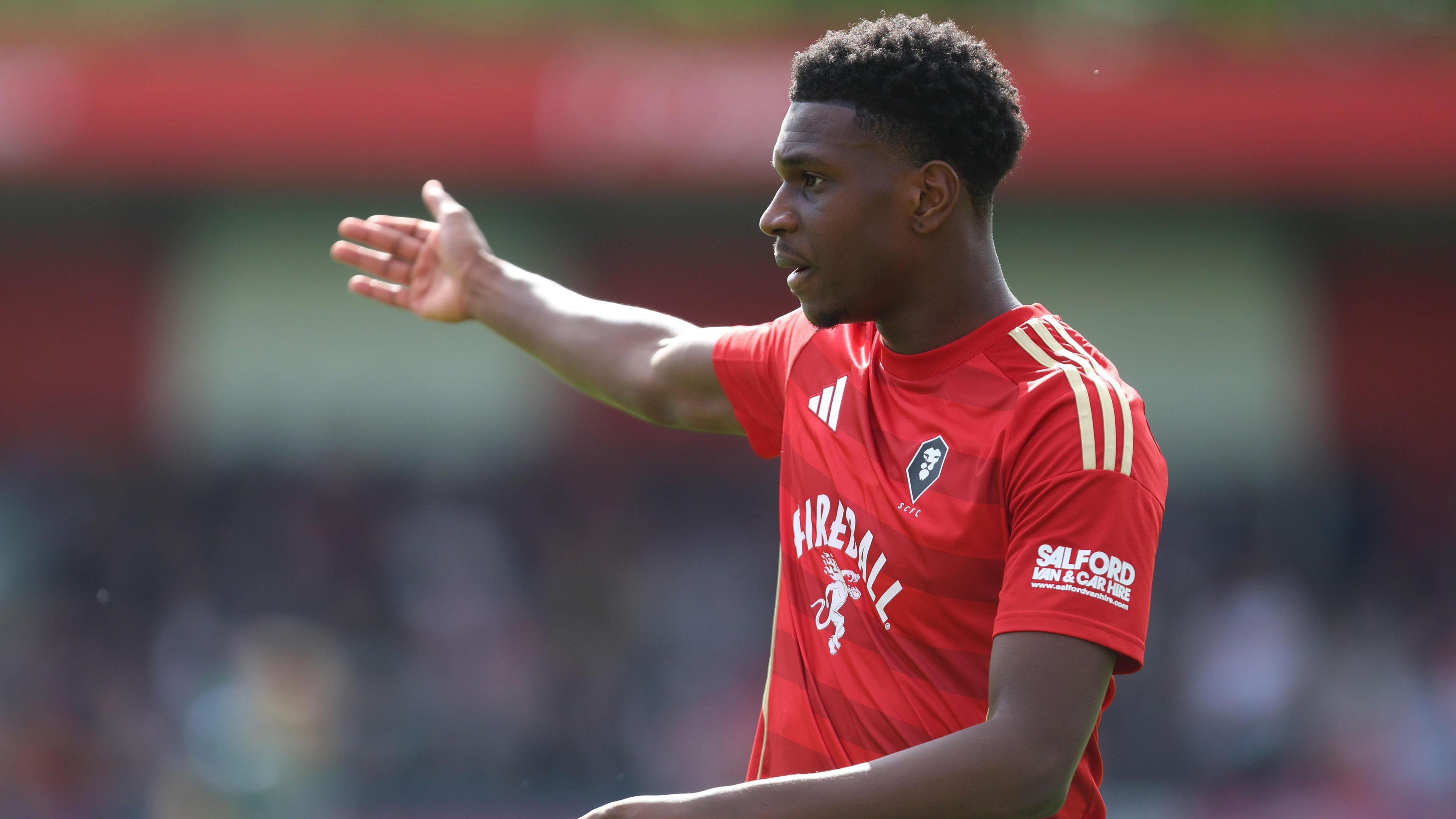 Salford City's Loick Ayina, wearing the club's red home shirt, orchestrates his defence with his right arm outstretched