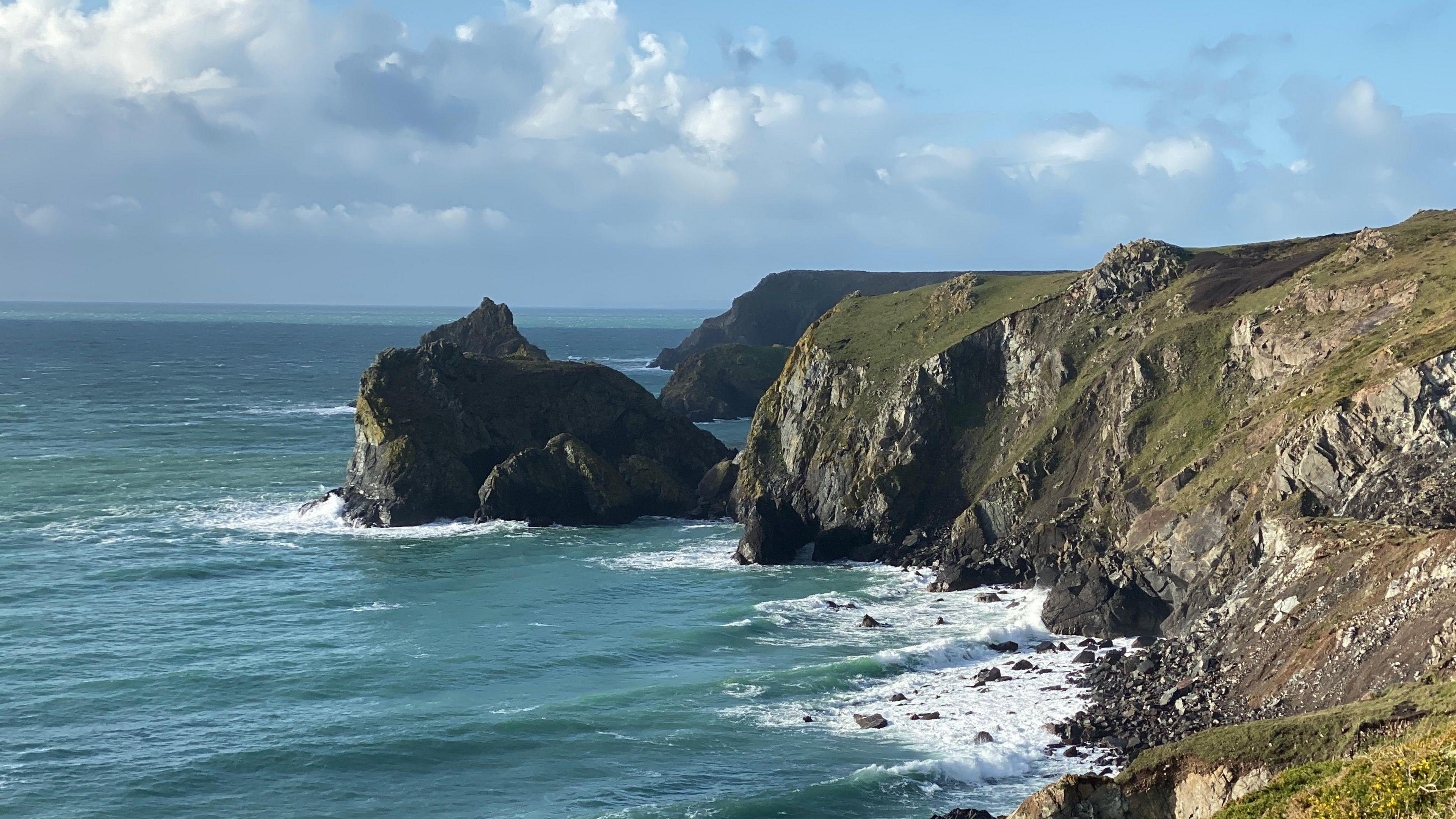 A shot of the coast near Lizard Point in Cornwall. On the left there is blue sea with a large rock in the distance. On the right is a cliff which is green and grey. There are waves crashing up against it. 