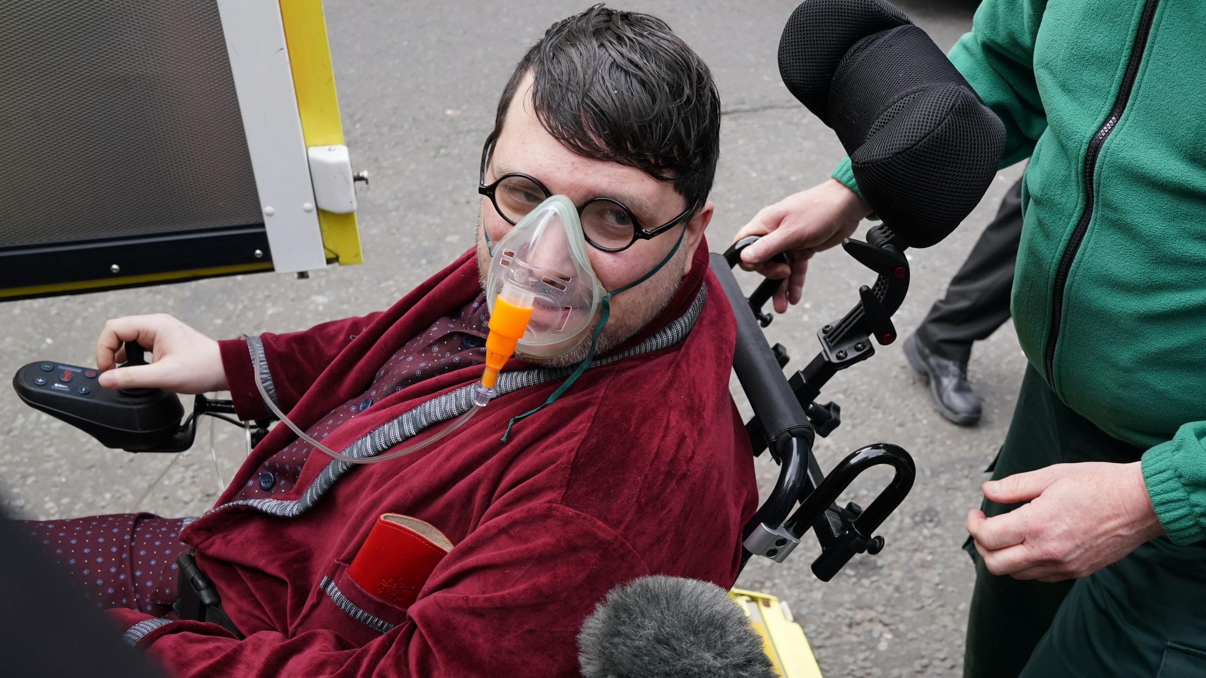 Nicholas Rossi outside court in a wheelchair, wearing a maroon dressing gown and silk pyjamas. He is wearing glasses and an oxygen mask.
