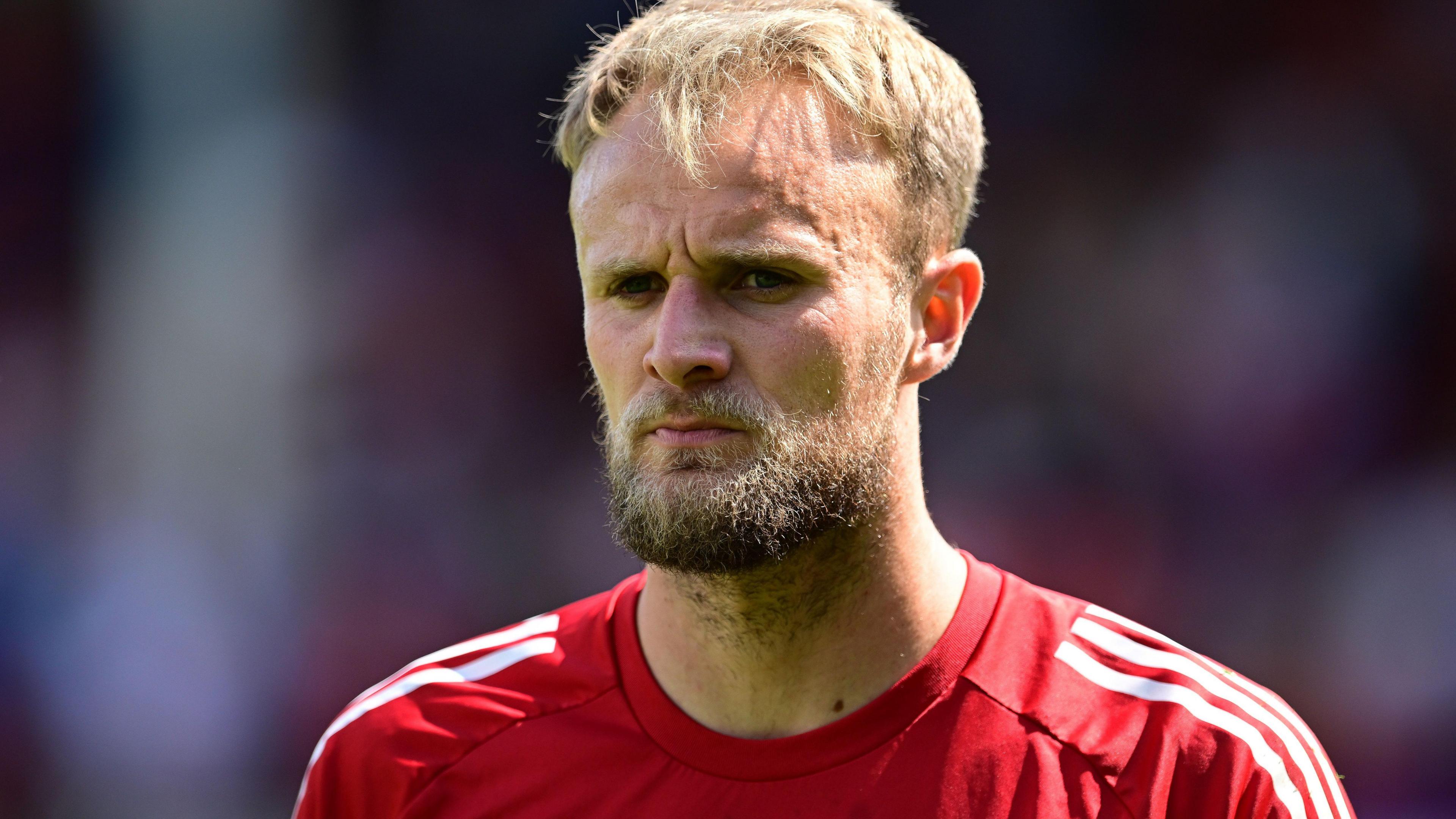 Lewis Ward is wearing a red goalkeepers top with white stripes on the shoulders. He has a blonde and brown beard.
