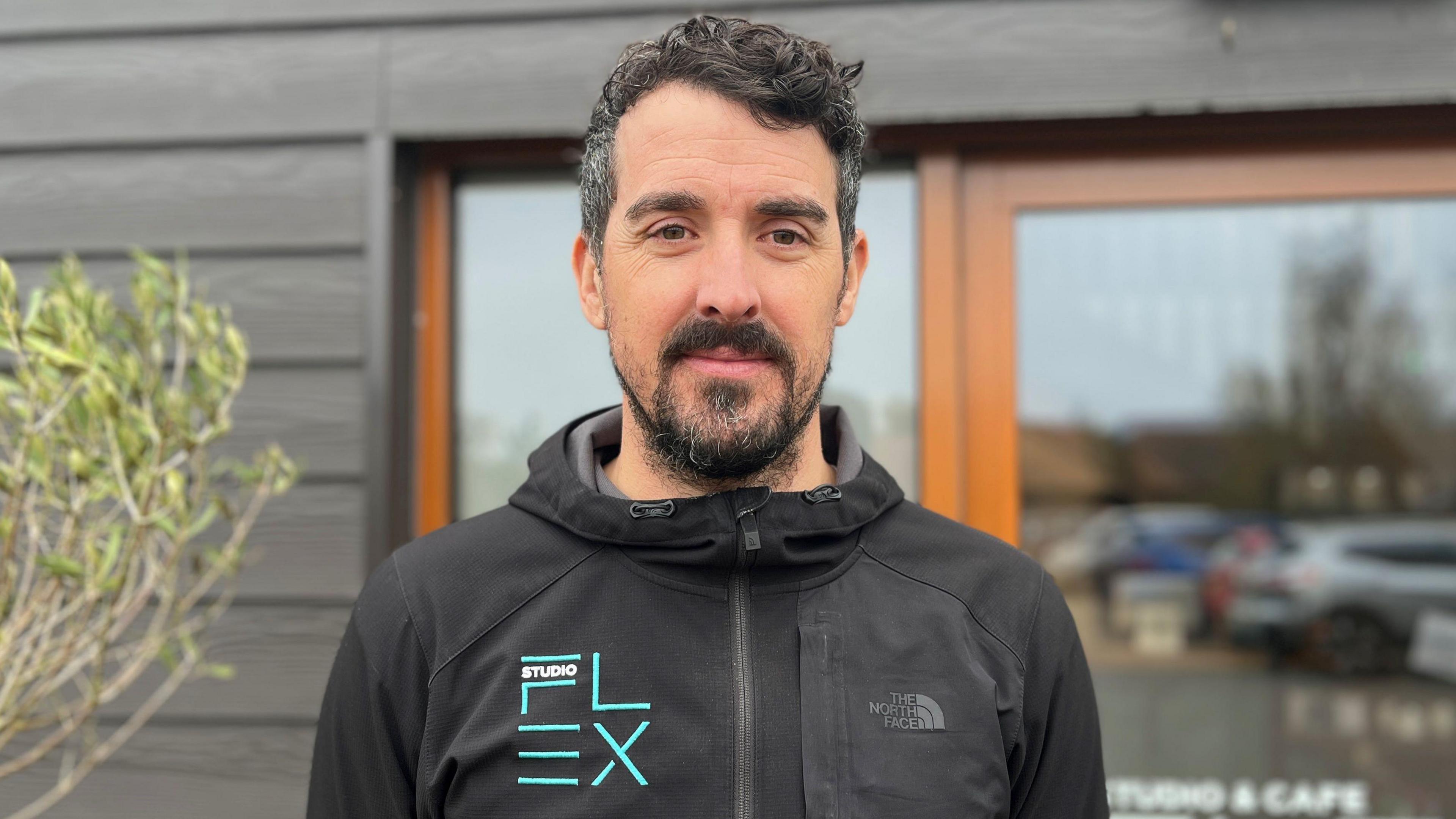 Head and shoulders of a 40-year-old man with short, dark hair with a beard and moustache.  He's wearing a black zip up top with Studio Flex branding. In the background is a glass doorway in a timber building. 