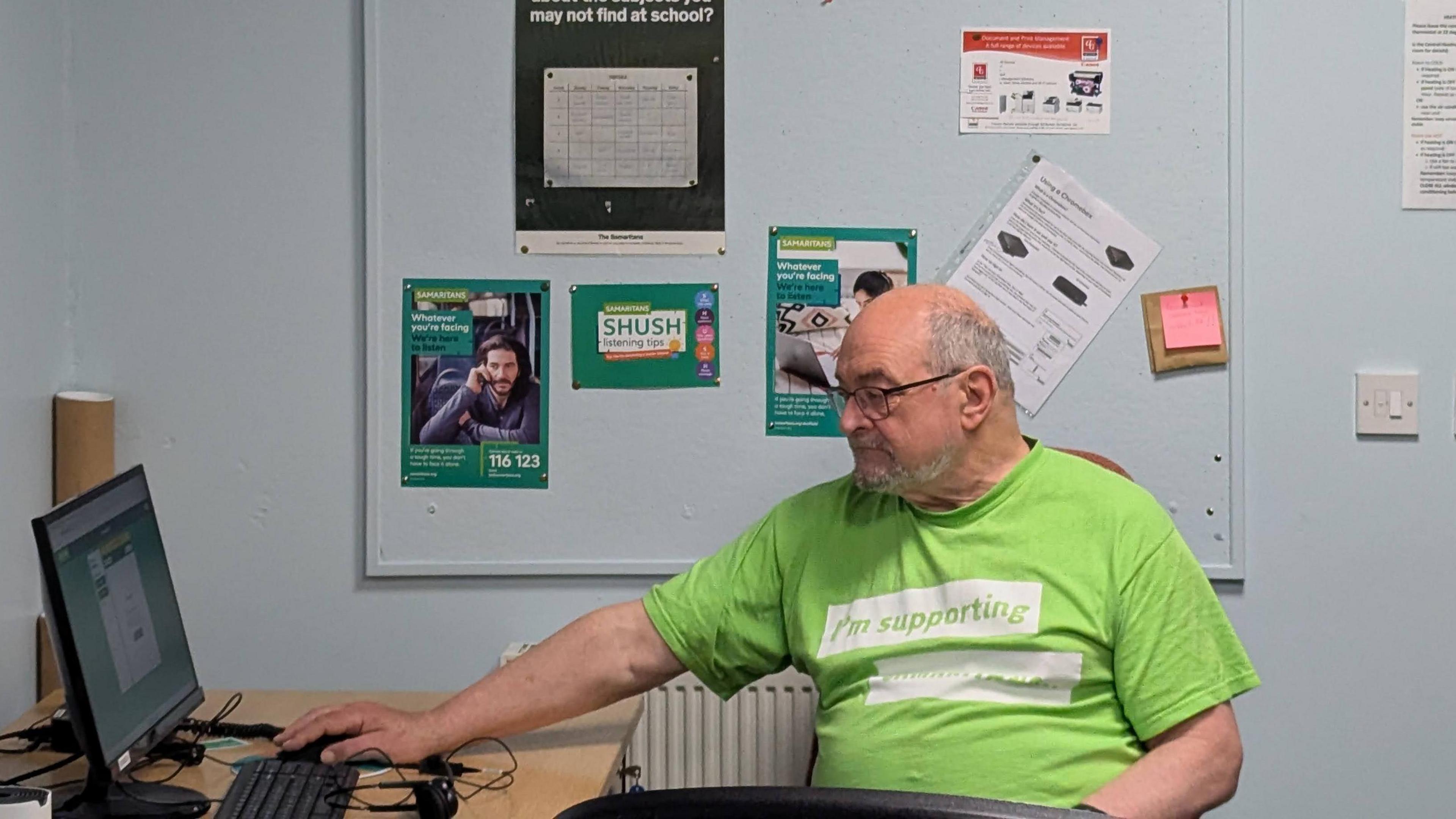 A man, Chris Collin, sits sideways looking at a computer screen on a desk, his hand on a pc mouse.