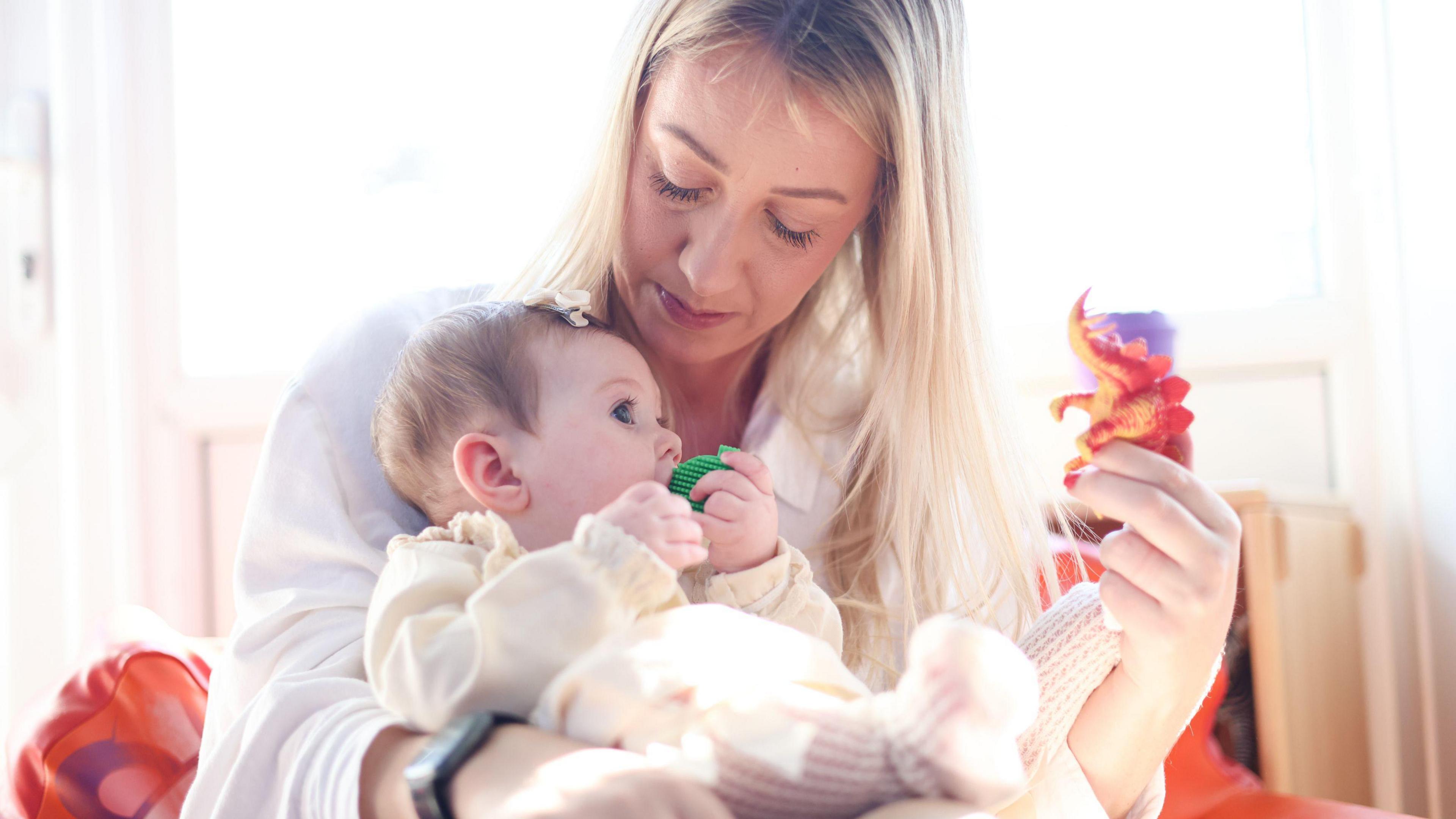 Hayley O'Sullivan holding one of her young daughters. Mrs O'Sullivan has long blonde hair and is wearing a white shirt. She is holding a red toy dinosaur in her left hand. Her baby is wearing a cream outfit and has a green dummy in her mouth.