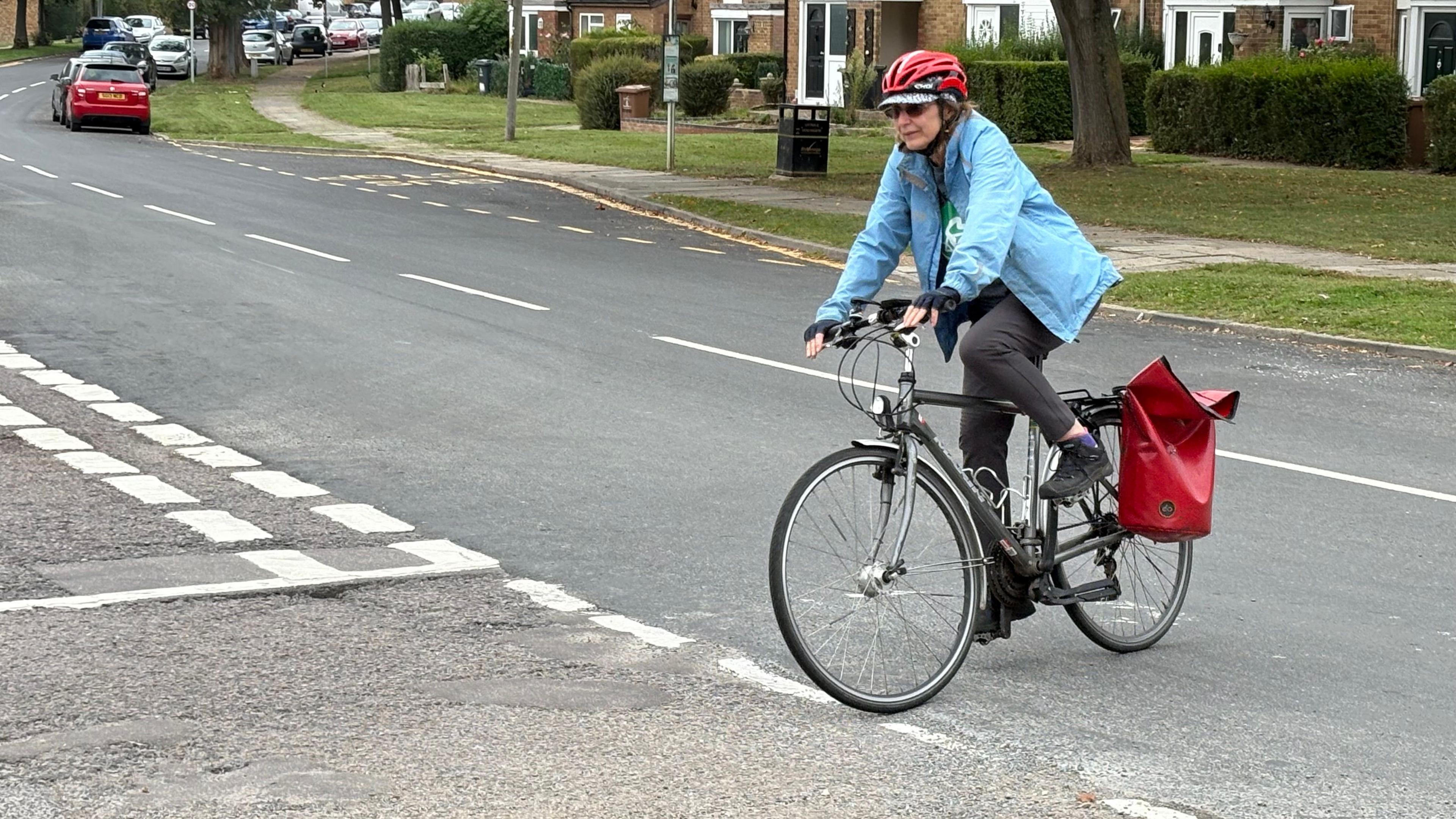 Tina Walker with red and white cycle helmet, light blue jacket and grey trousers, cycling from a main road into a side road with a red bag on the back of her bike.There is a path with grass either side behind her. Parked cars can be seen in the distance.