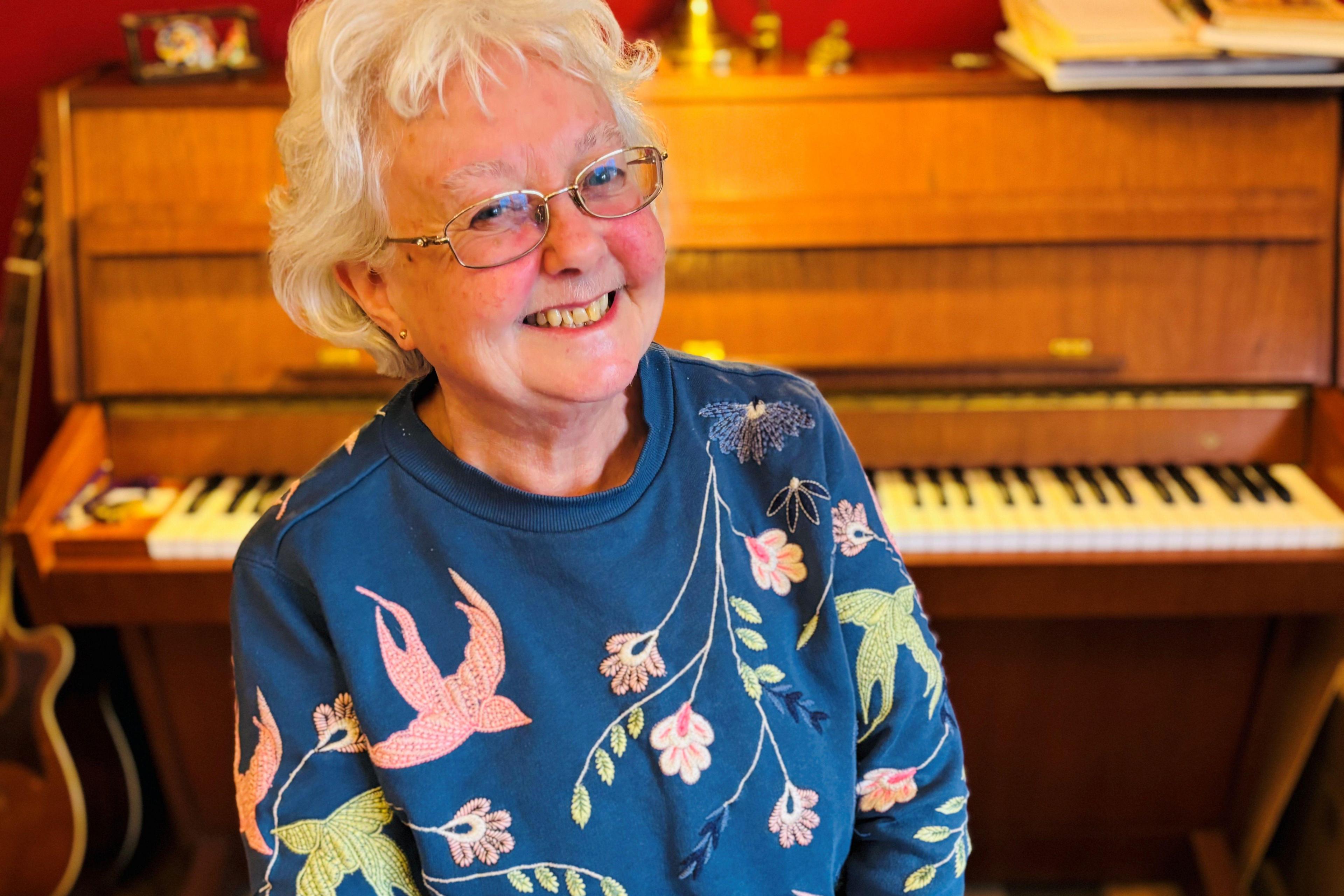 Liz Slaven, who has grey hair and glasses, smiling at camera, with a piano in the background.