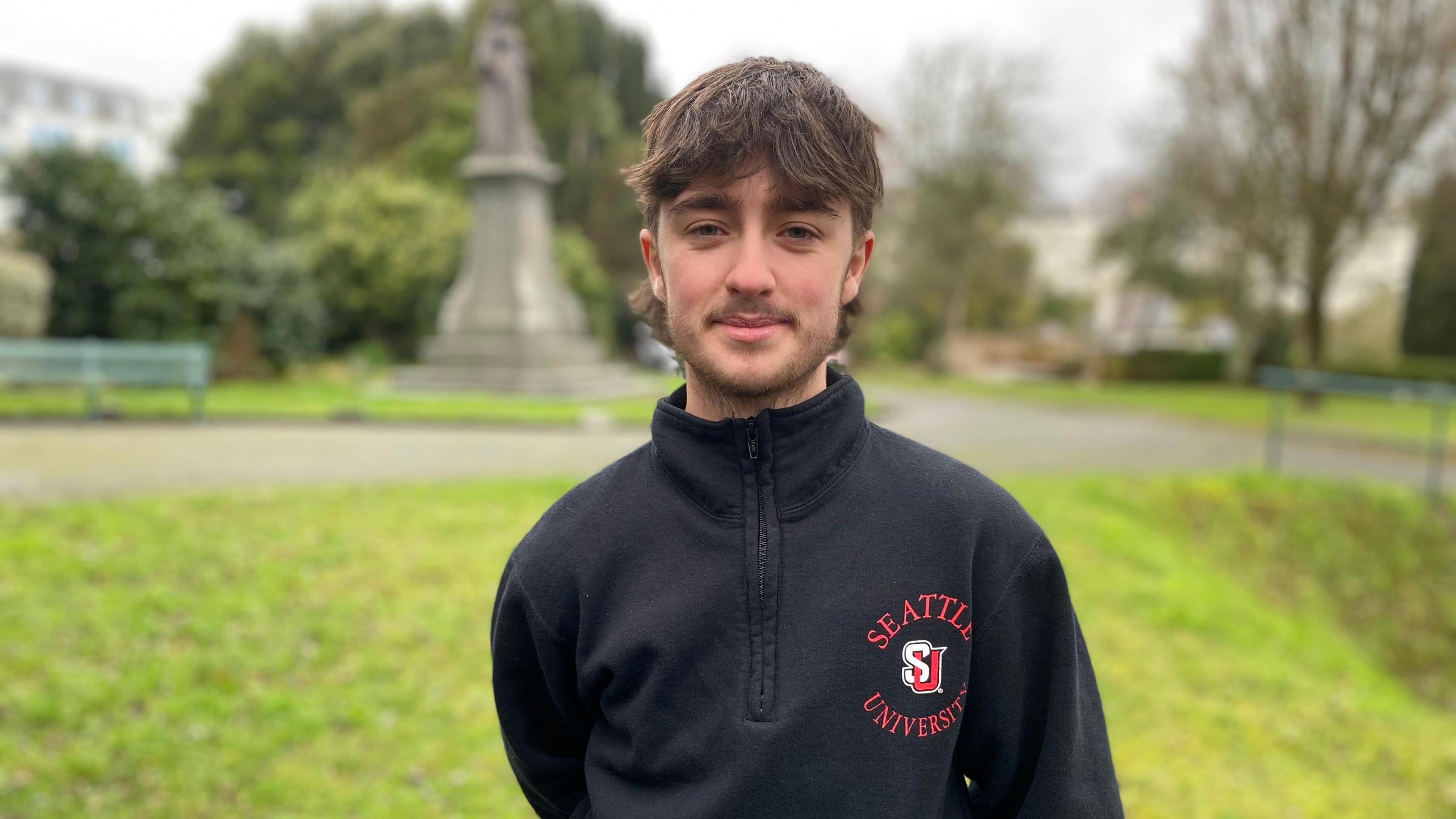 A young man with dark brown hair and a short beard is wearing a black jumper.