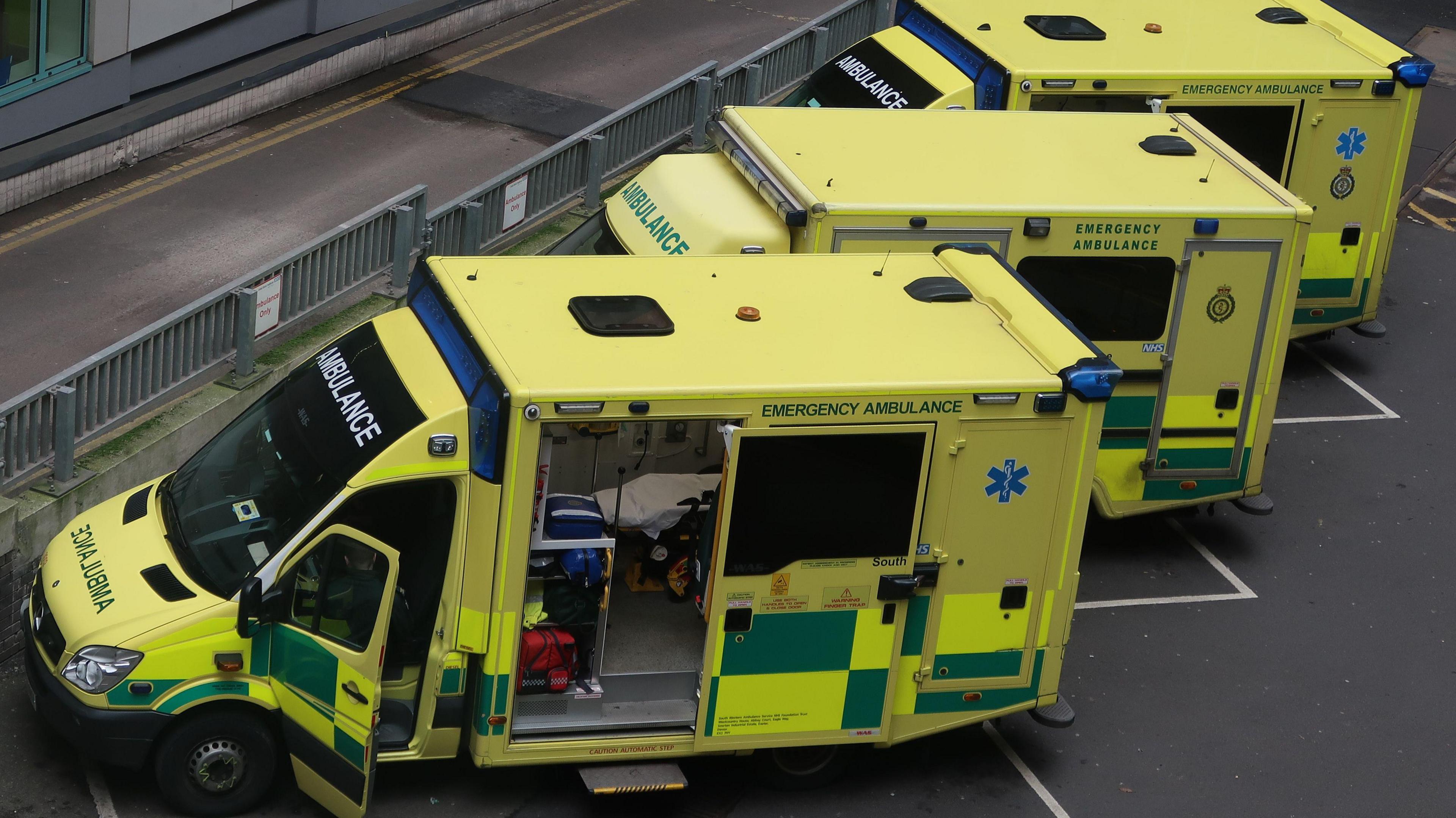 Three ambulances parked up outside a hospital. One of them has the side door open and a bed and medical equipment can be seen inside.