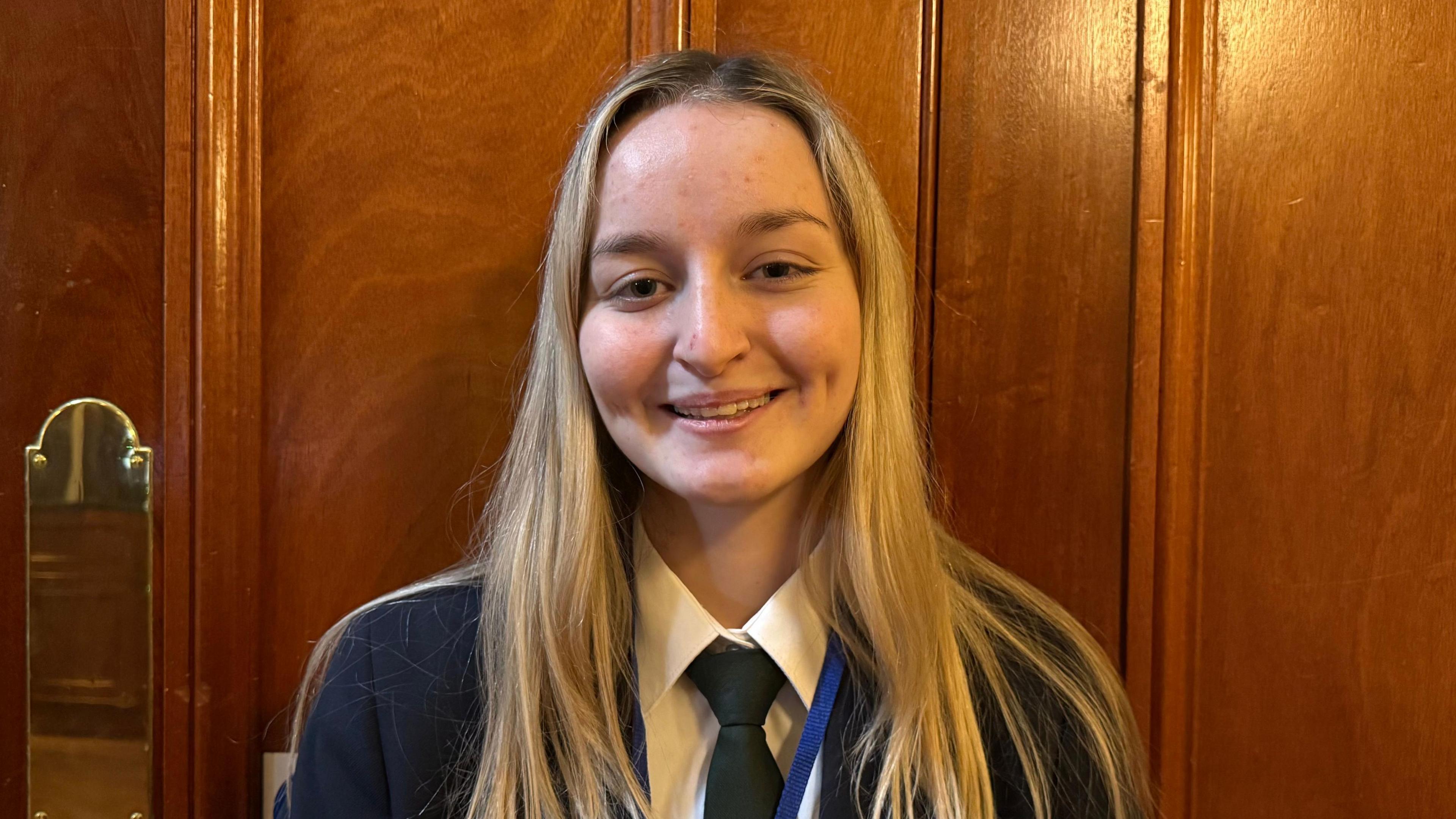 A teenage girl with blonde hair, wearing a dark blazer, white shirt and dark green tie, smiling at the camera. She is standing in front of a brown wooden door.