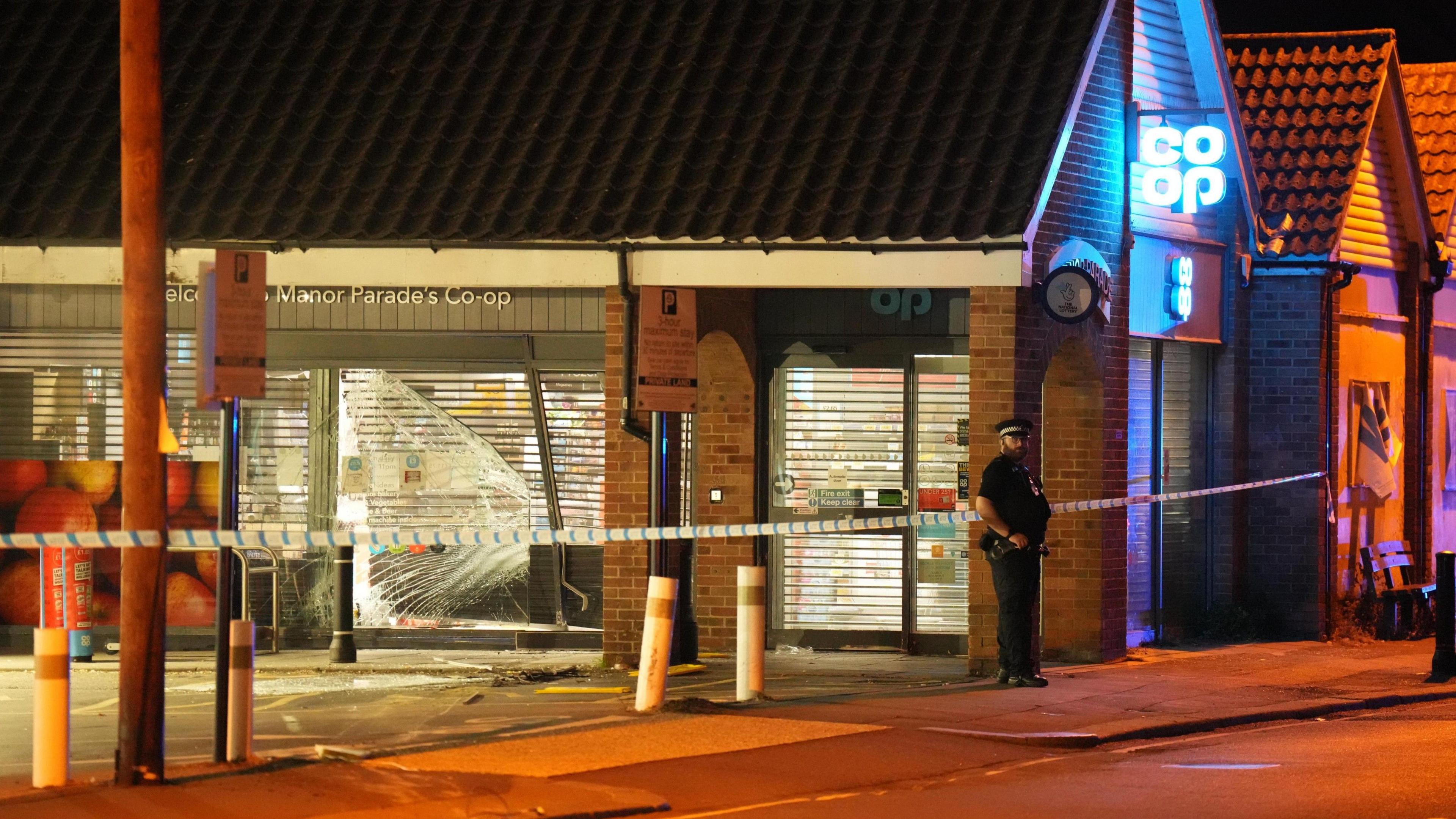 An exterior view of a parade of shops in Worthing that have been ram raided by a vehicle. The metal shutter of a supermarket has been smashed in, as has the glass frontage of the store, while a concrete bollard is at an angle. A lone police officer stands guard at the scene.