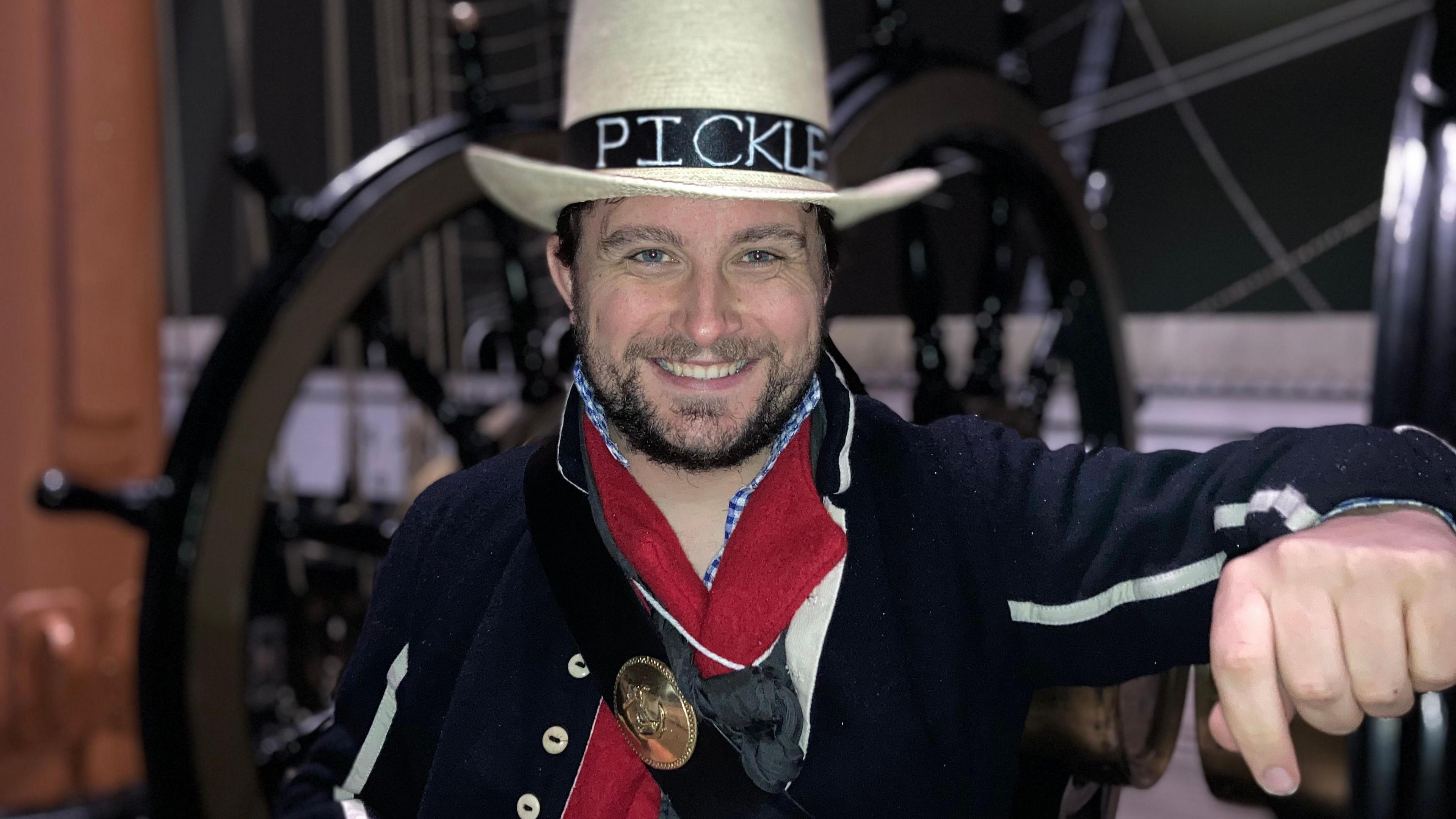 Man standing in traditional 19th Century seaman dress, leaning against a large wheel on HMS Warrior