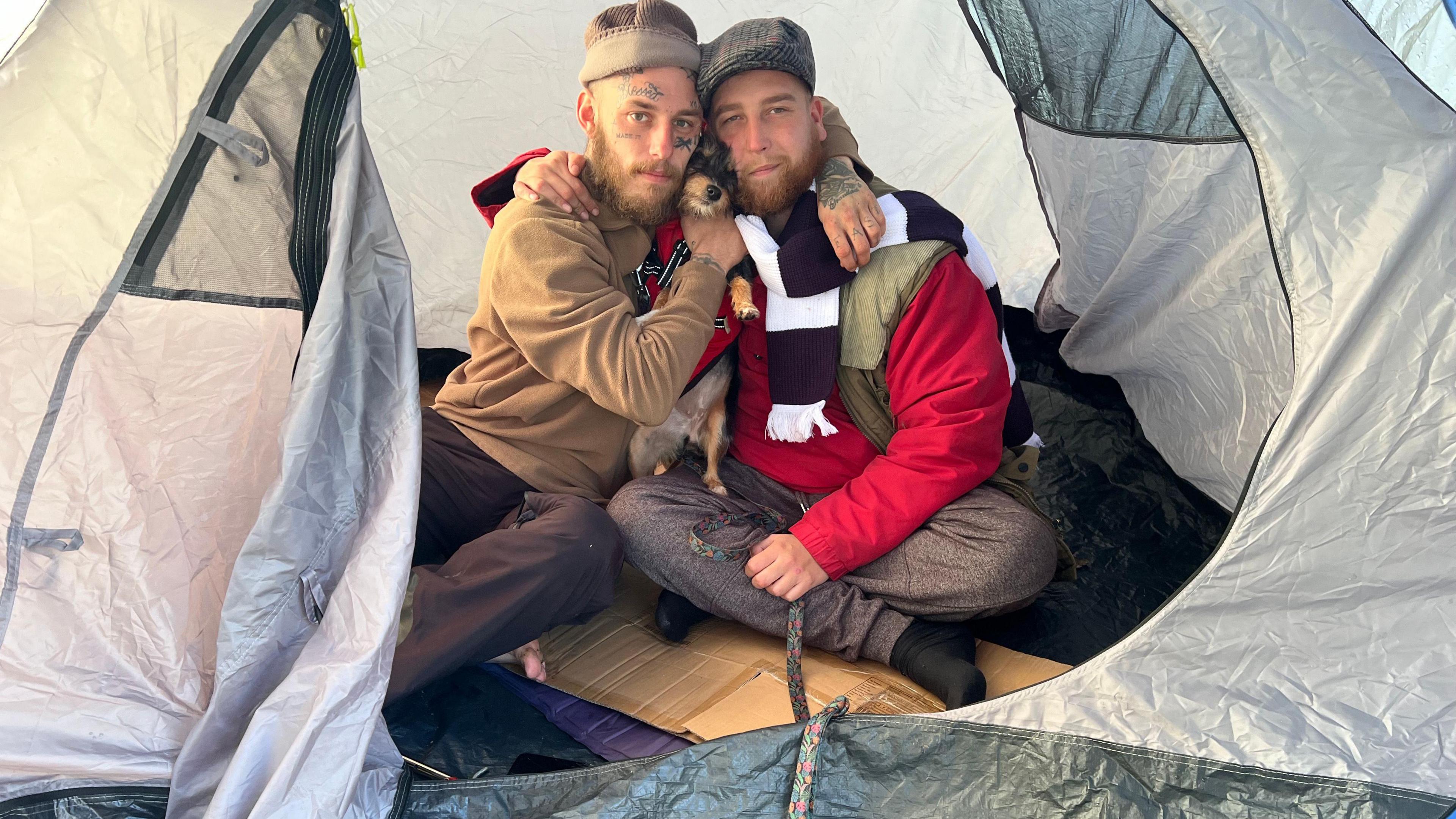 Two men with wearing hats are seen sitting inside a tent, cuddling a small dog.